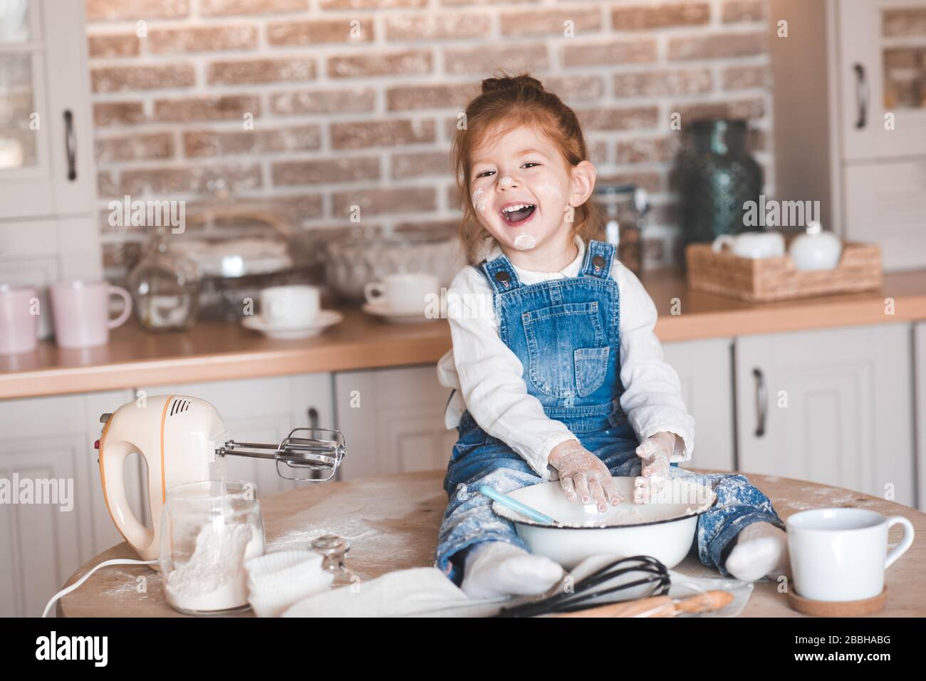 Das lachende Mädchen, das 3-4 Jahre alt ist und Gebäck auf dem Tisch in der Küchenecke sitzt. Blick auf die Kamera. Kindheit. Stockfoto