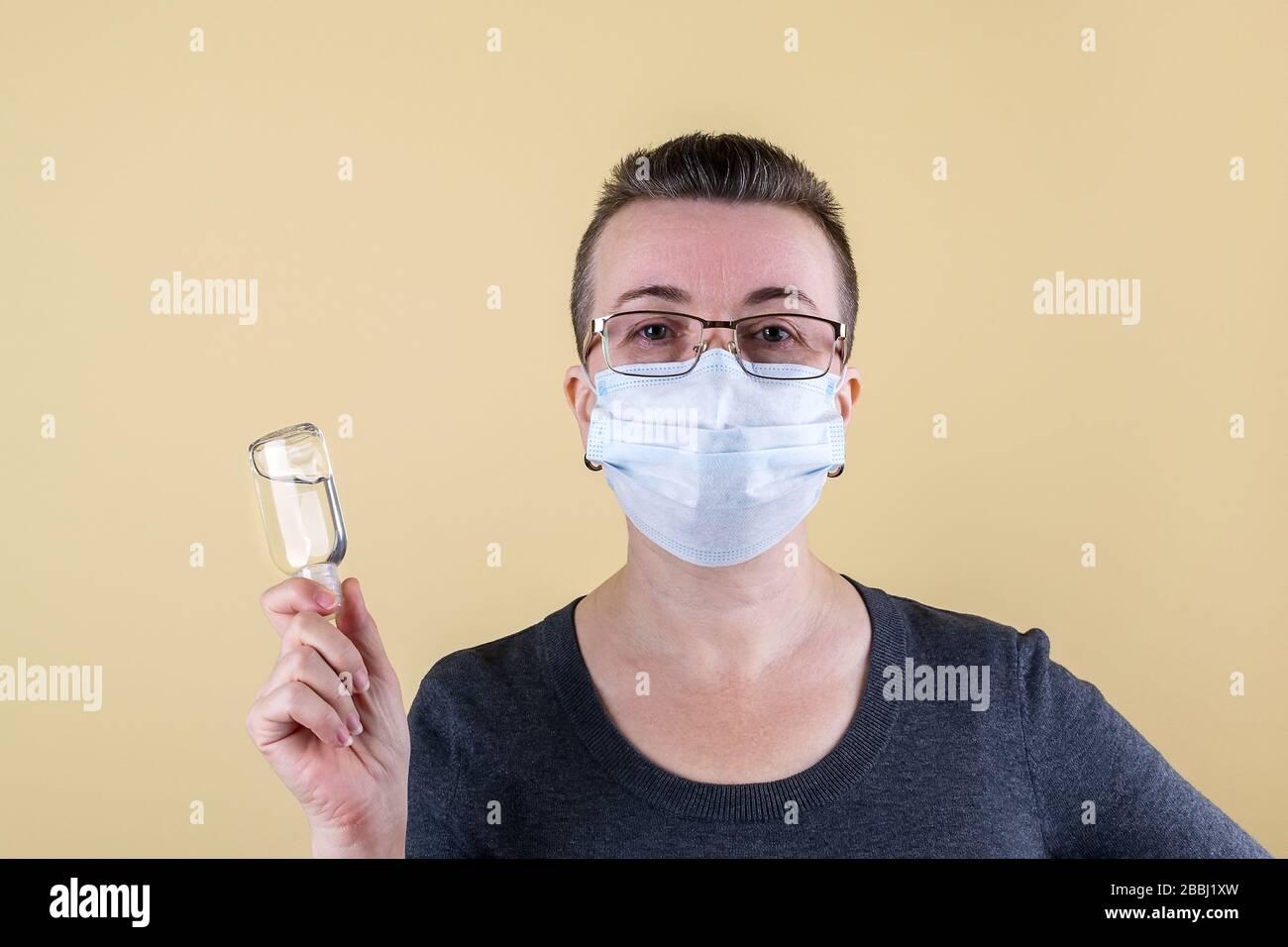 Sehr kurzhaarige kaukasische Erwachsene Frau im Pullover und mit Brille und medizinischer Maske auf dem Gesicht hält einen Krug mit antiseptischem Gel in der Hand. Stockfoto