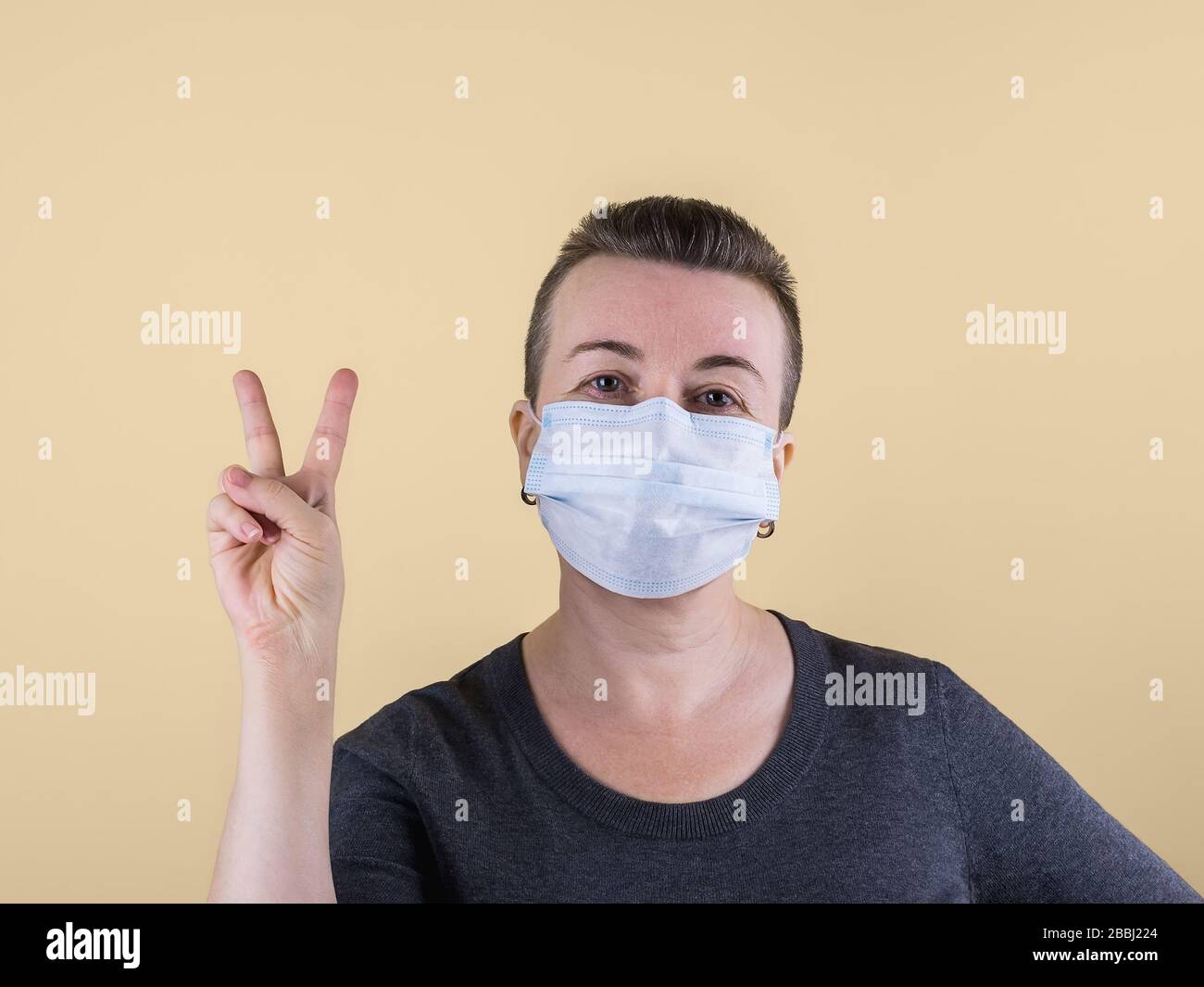 Sehr kurzhaarige kaukasische Erwachsene Frau im Pullover und mit medizinischer Maske auf dem Gesicht zeigt Siegeshandgeste. Auf beigefarbenem Hintergrund. Kopierbereich. Stockfoto