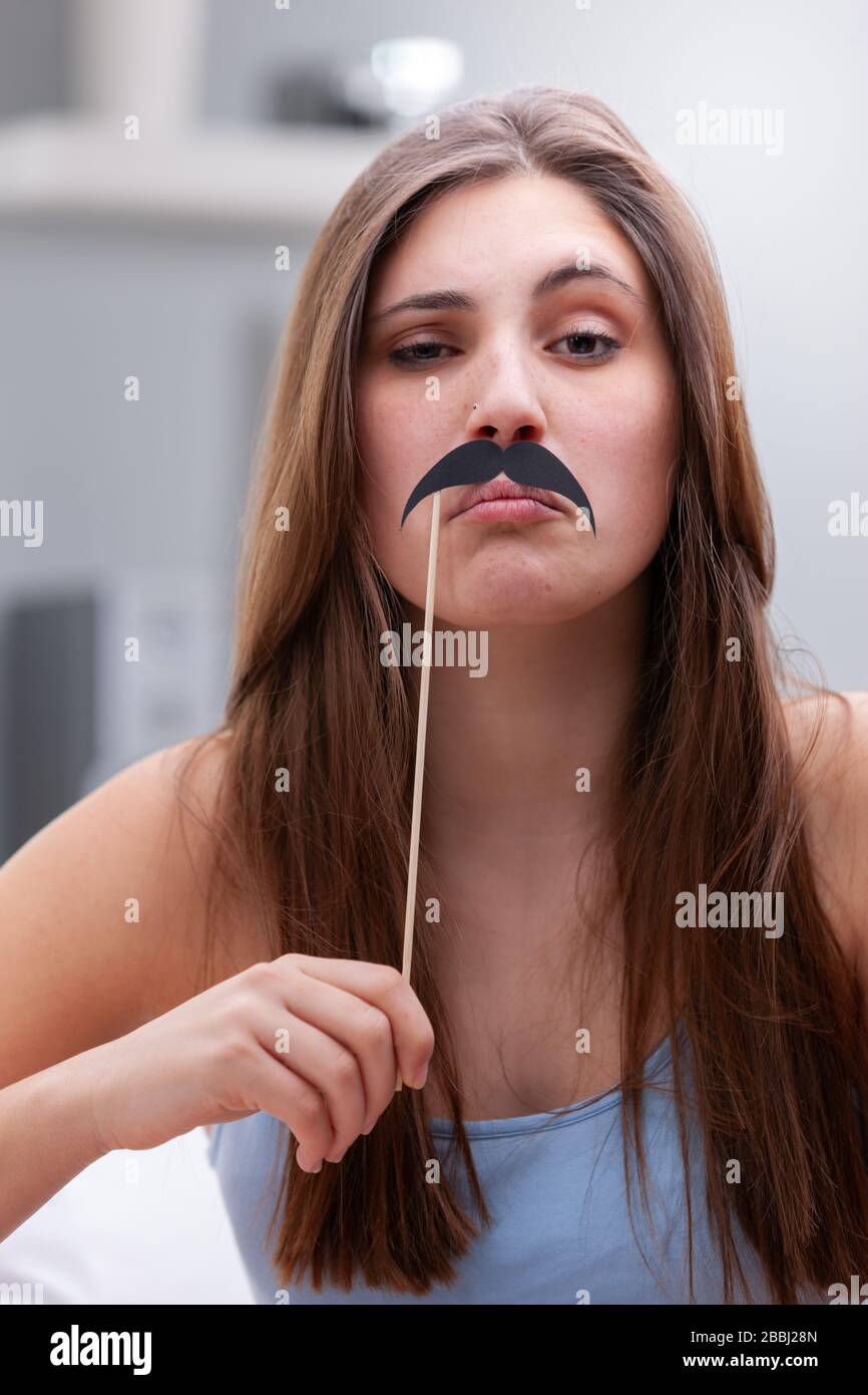 Junges Mädchen im Teenager-Alter, das mit Party-Accessoires umhergeht, die einen Schnurrbart mit einem überhöhlichen Ausdruck an der Oberlippe halten, in geschlossenen Räumen Stockfoto