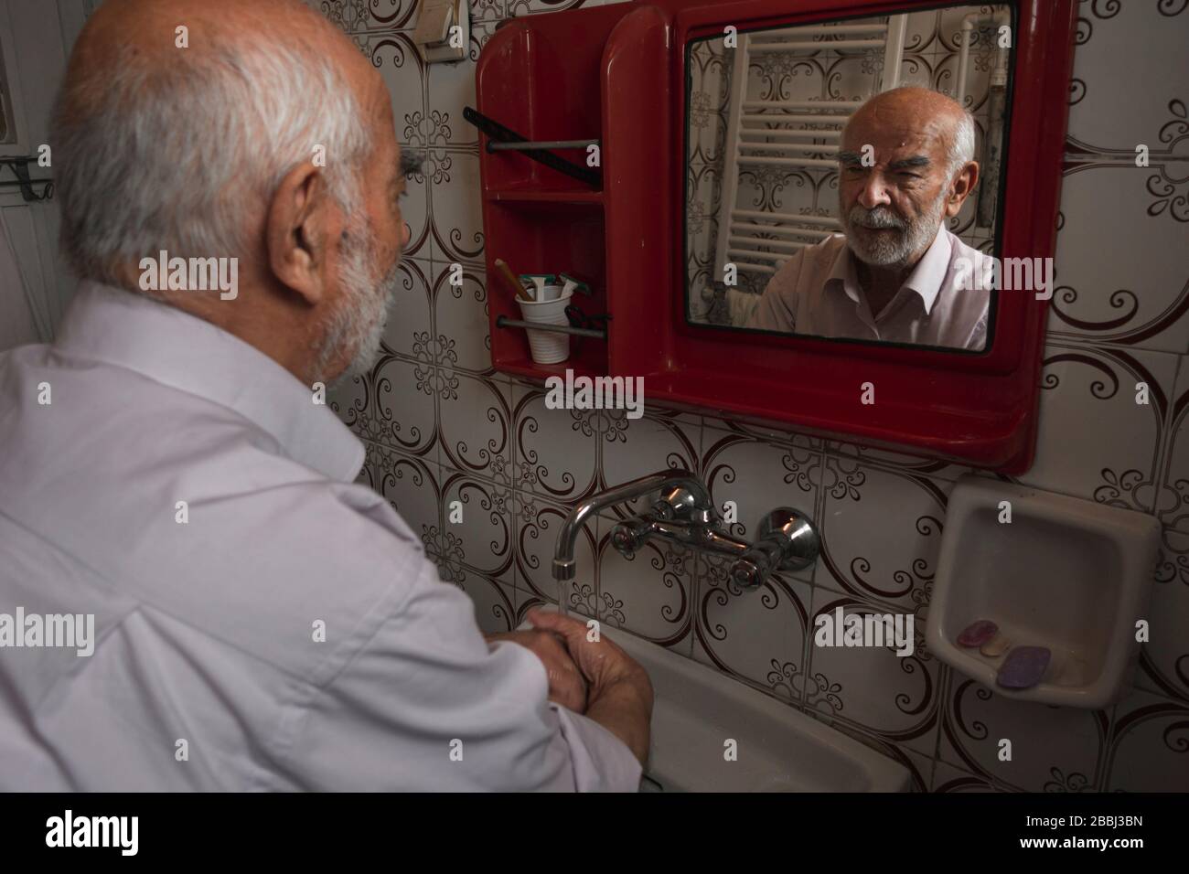 80 Jahre alter türkischer Mann, der in seinem alten Badezimmer Spiegel schaut, während er seine Hände waschen Stockfoto