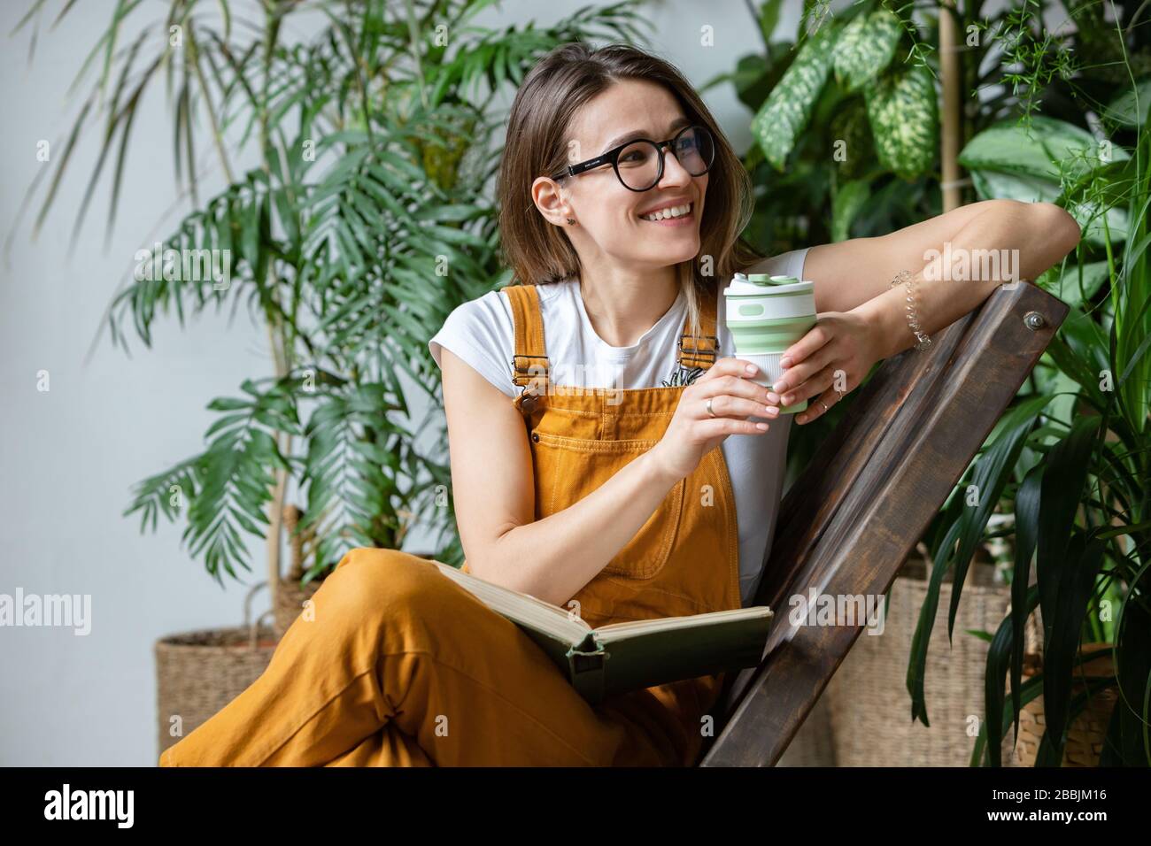 Weibliche europäische Gärtnerin, die Overalls trägt, nach der Arbeit ruht, auf Holzstuhl im heimischen Gewächshaus sitzt, wiederverwendbaren Kaffee-/Teebecher beiseite hält Stockfoto