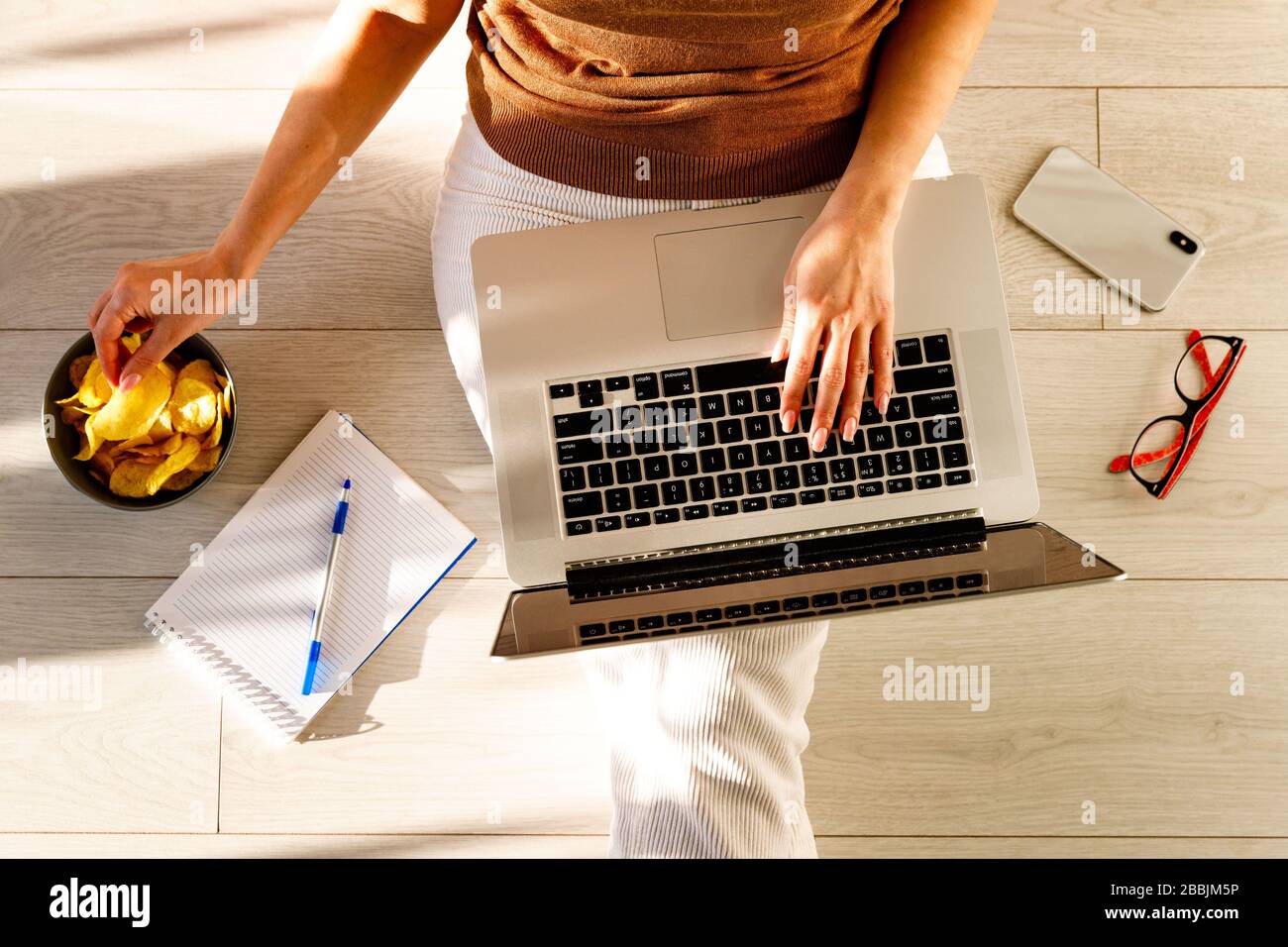 Flaches Lay von Frau, die drinnen auf dem Boden sitzt, berührt und isst Pommes. Smartphone, Brille, Notebook liegen in der Nähe. Gemächliche Remotearbeiten auf Laptop duri Stockfoto
