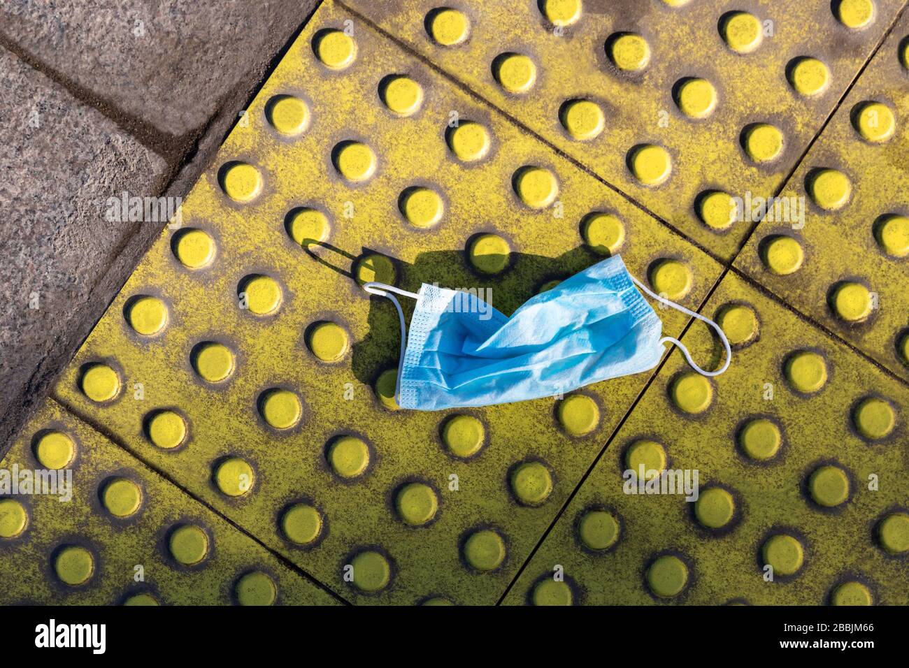 Blau schmutzig verwendete chirurgische Gesichtsmaske zum Schutz von Coronavirus auf gelben taktilen Pflastersteinen. Entsorgung medizinischer Abfälle. Draufsicht. Allergiesaison, Staubluft. Stockfoto