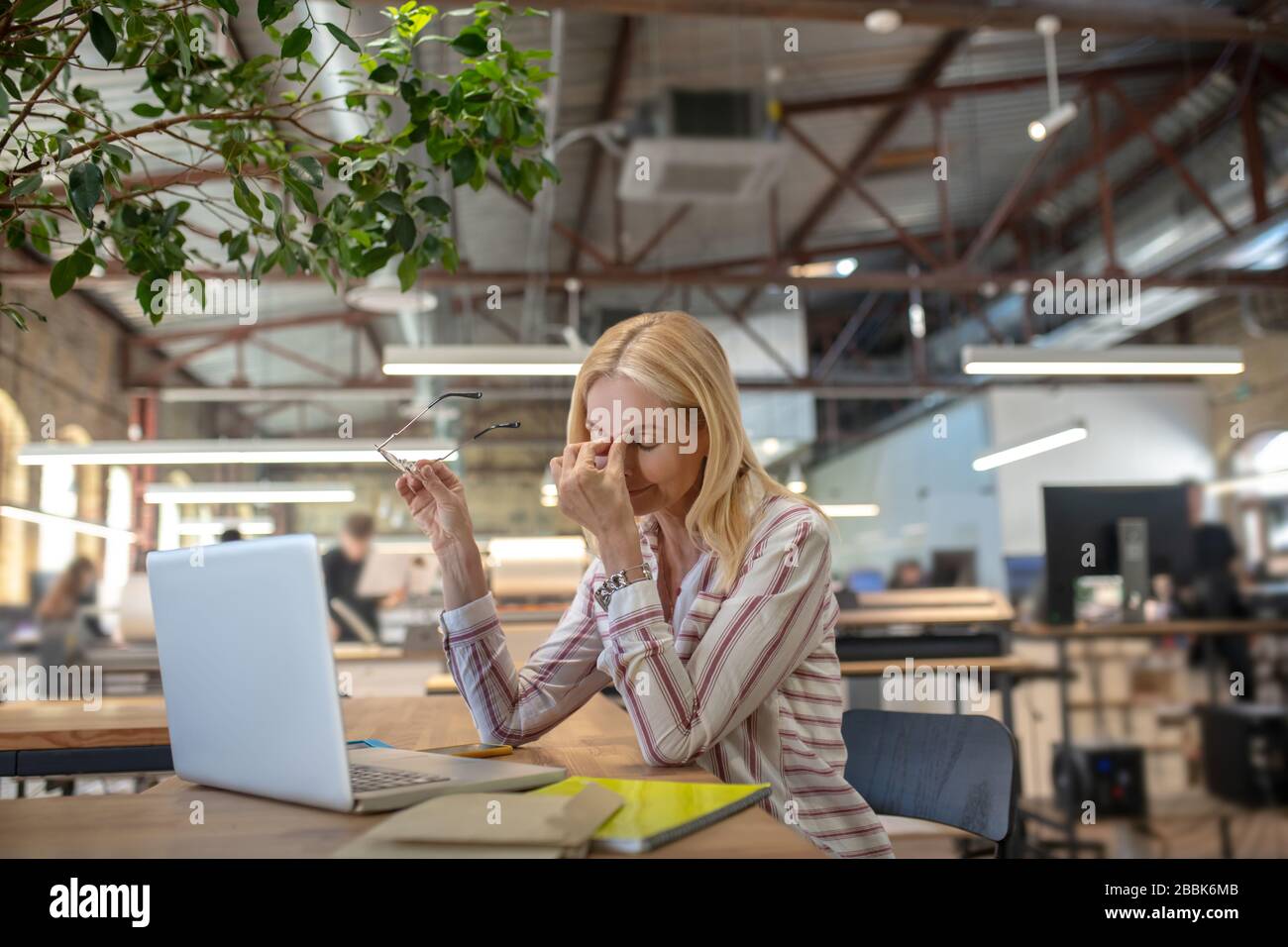 Blonde Frau sitzt am Laptop, massiert ihre Nasenbrücke und hält Brillen Stockfoto