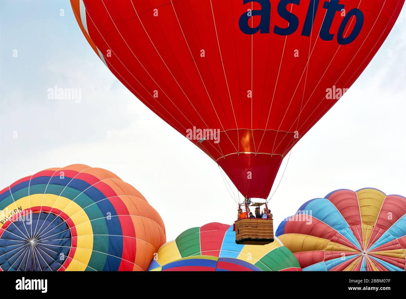 Roter Heißluftballon mit Korb und Menschen, die während des 34. Friese Ballonfeesten Festivals in Joure, Niederlande, über bunten Ballons steigen. Stockfoto