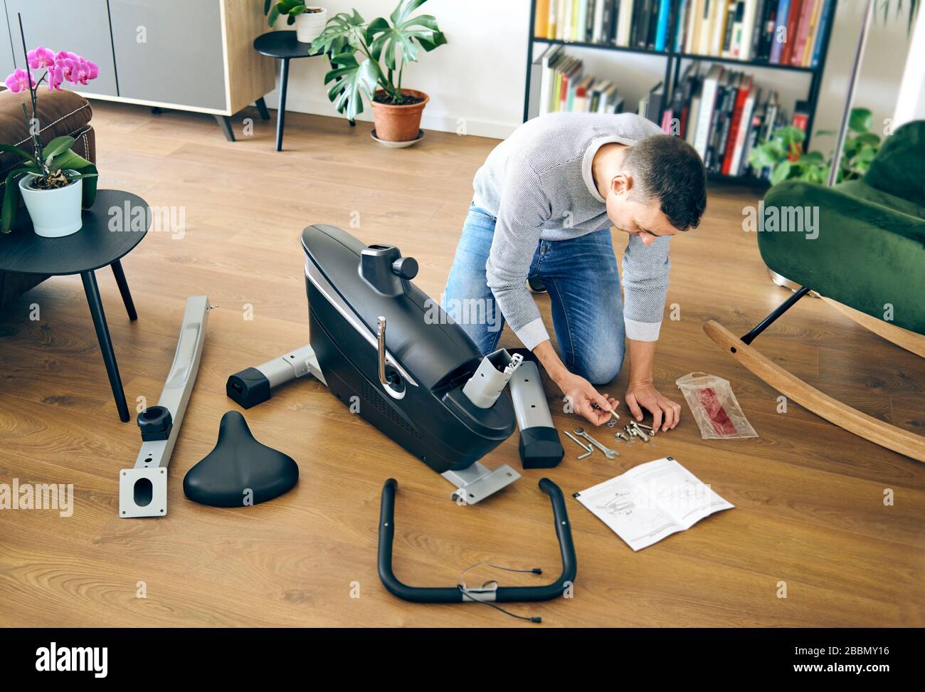 Ein kaukasischer Mann, der die verschiedenen Teile eines stationären Fahrrads im Wohnzimmer seines Hauses zusammenstellte Stockfoto