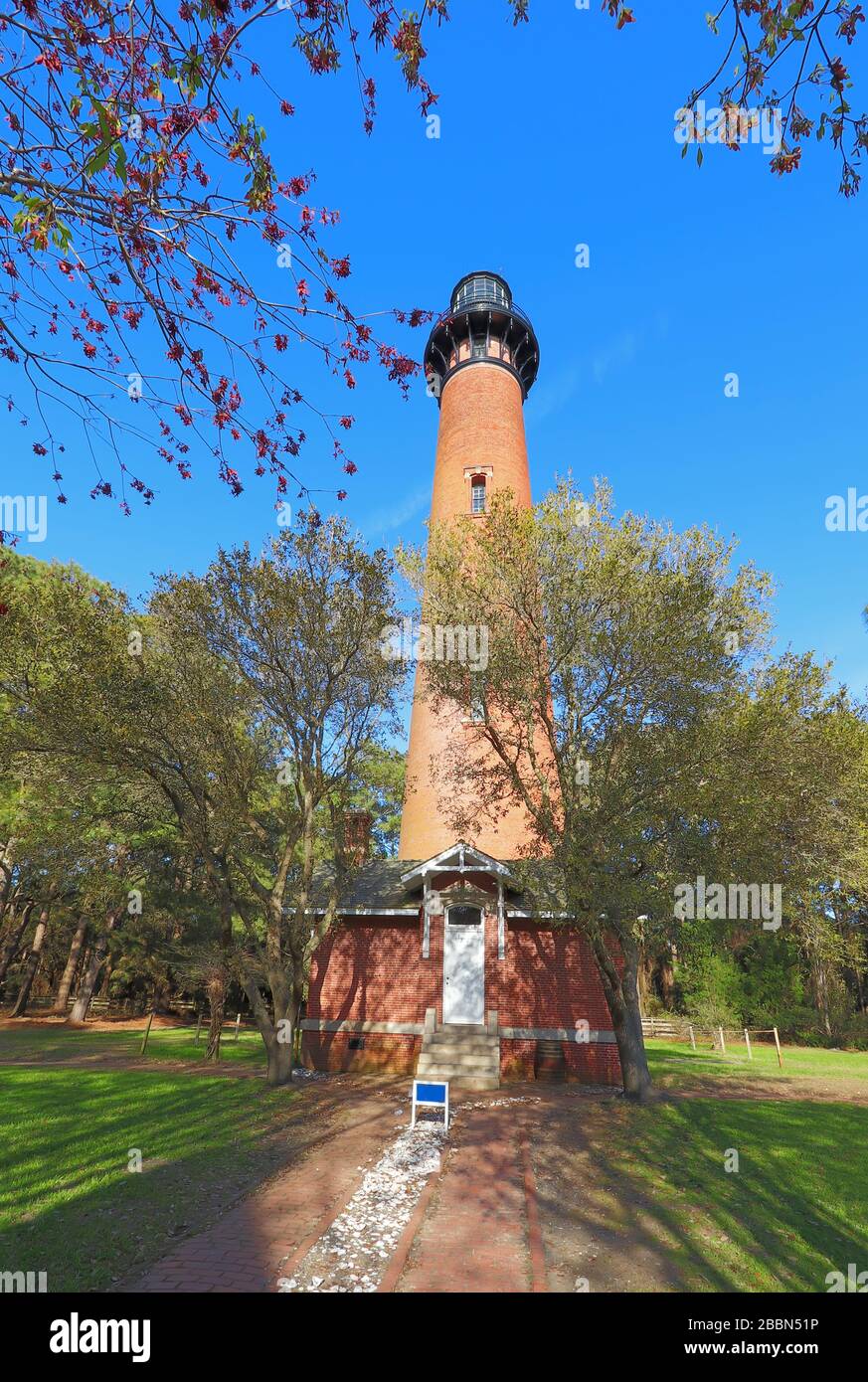 Die rote Ziegelsteinstruktur des Currituck Beach Lighthouse ragt über ein kleines Einsteigerhaus und Kiefern im Currituck Heritage Park in der Nähe von Corolla, N Stockfoto