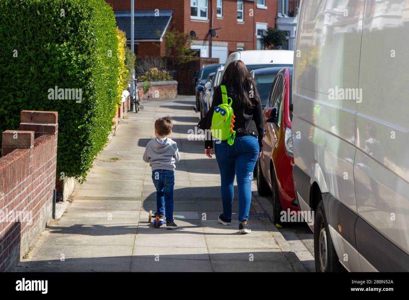 Eine Mutter, die neben einem kleinen Kind spazieren geht, das einen Roller fährt Stockfoto