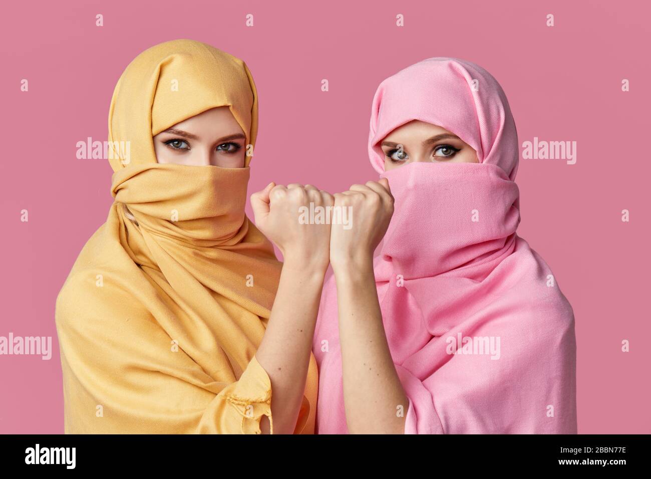 Mädchen Macht. Zwei junge muslimische araberinnen tragen bunte Hijabs vor pinkfarbenem Hintergrund. Zeigt den Finger zur Seite. Leerzeichen für Text Stockfoto