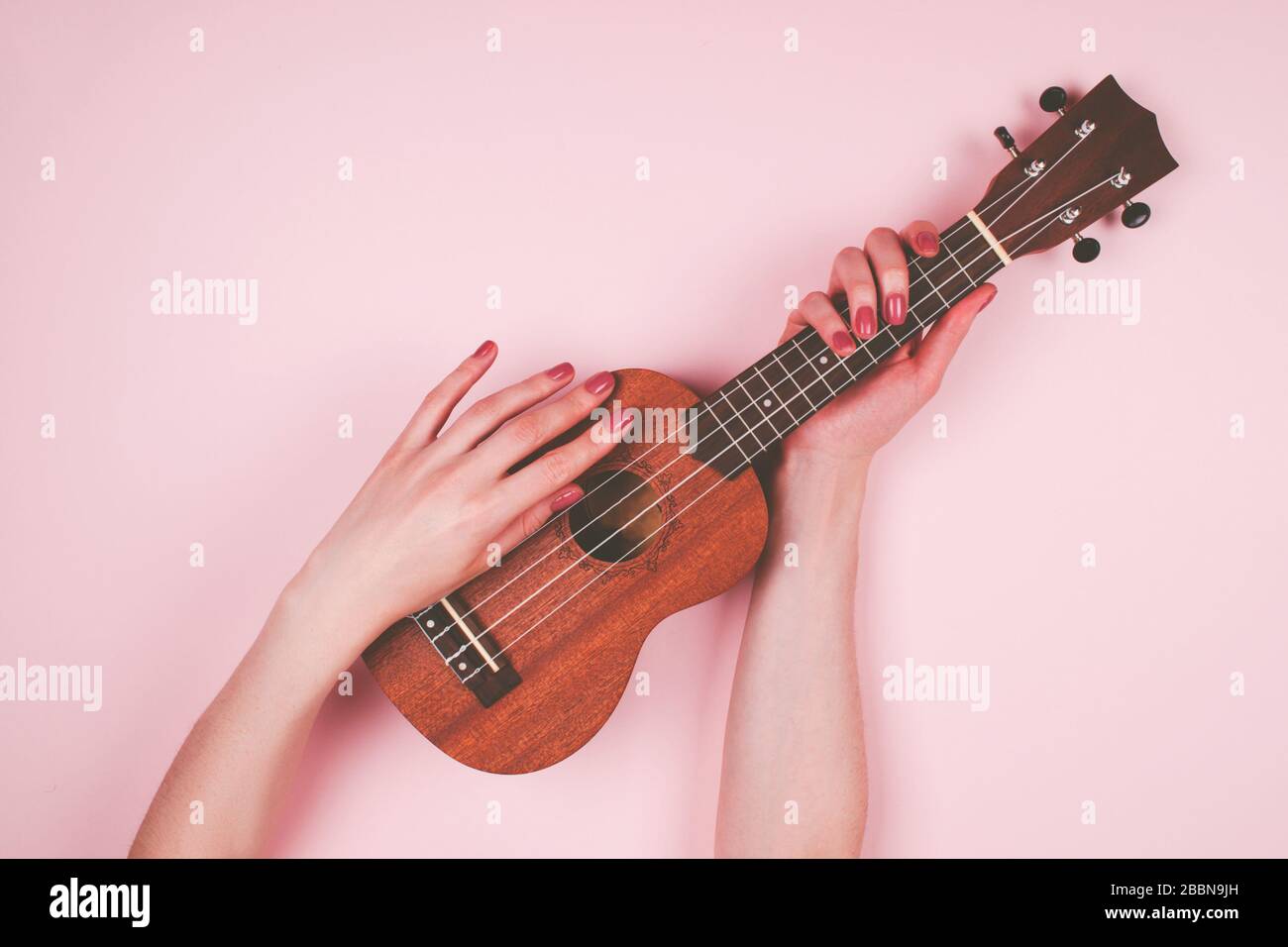 Frau Hände mit pinkfarbener Maniküre, die wenig Wodden Ukulele auf pinkfarbenem Hintergrund hält. Feines Musikerbild. Stockfoto