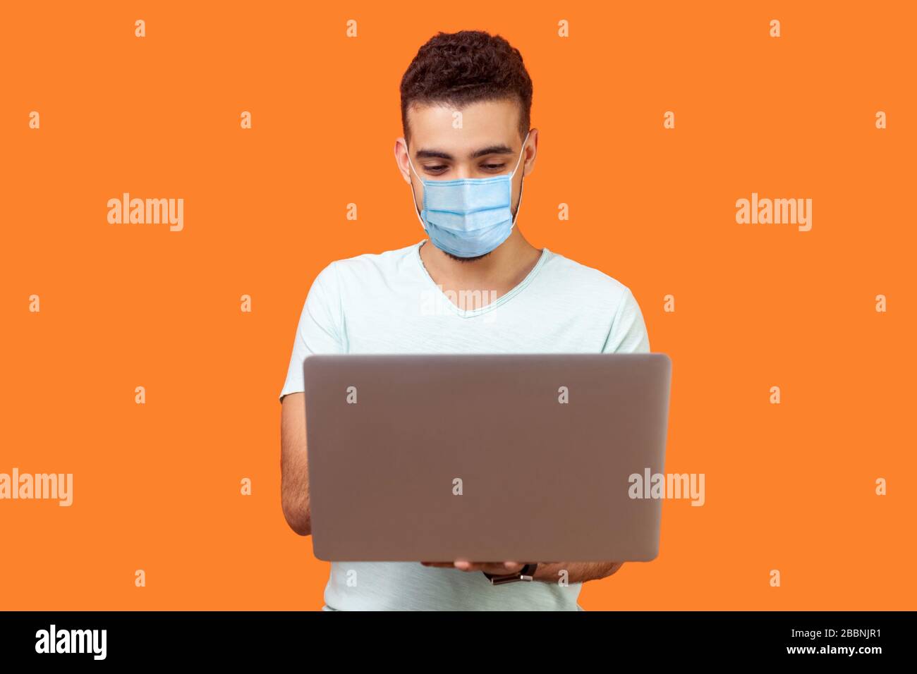 Portrait des fröhlichen jungen brünette Mannes mit chirurgischer medizinischer Maske, die E-Mails auf dem Laptop tippt und lächelt, positive Nachrichten liest oder im Internet surft. In Stockfoto