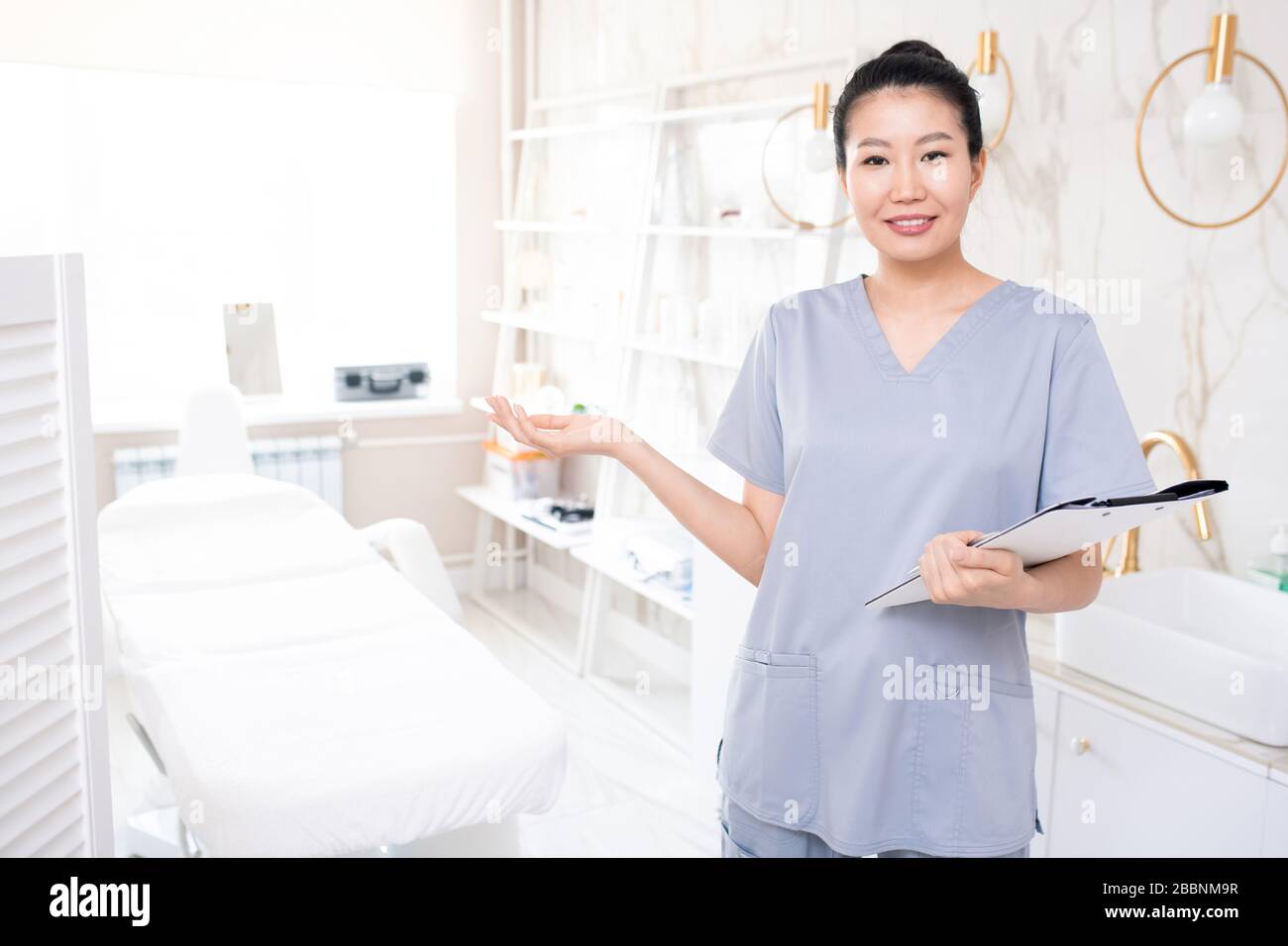 Portrait des lächelnden asiatischen Kosmetologen in den mit Zwischenablage stehenden Schruben und lädt Sie ein, Hautpflegeverfahren durchzuführen Stockfoto
