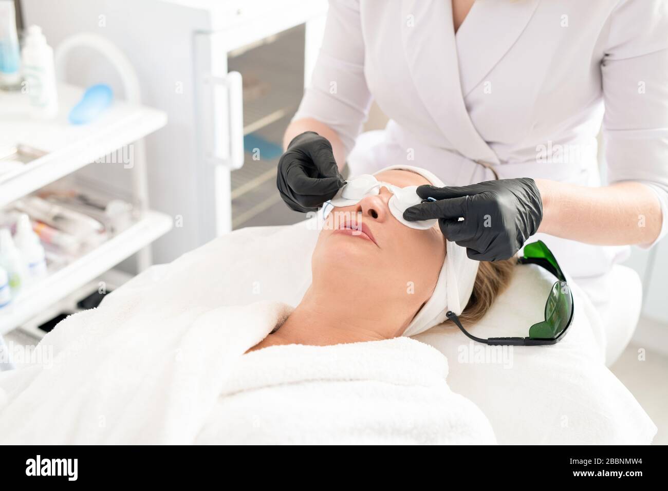 Nahaufnahme der nicht erkennbaren Kosmetikerin, die eine Laserschutzbrille auf das Gesicht der Frauen stellt, während der Kunde für die Laserchirurgie vorbereitet wird Stockfoto