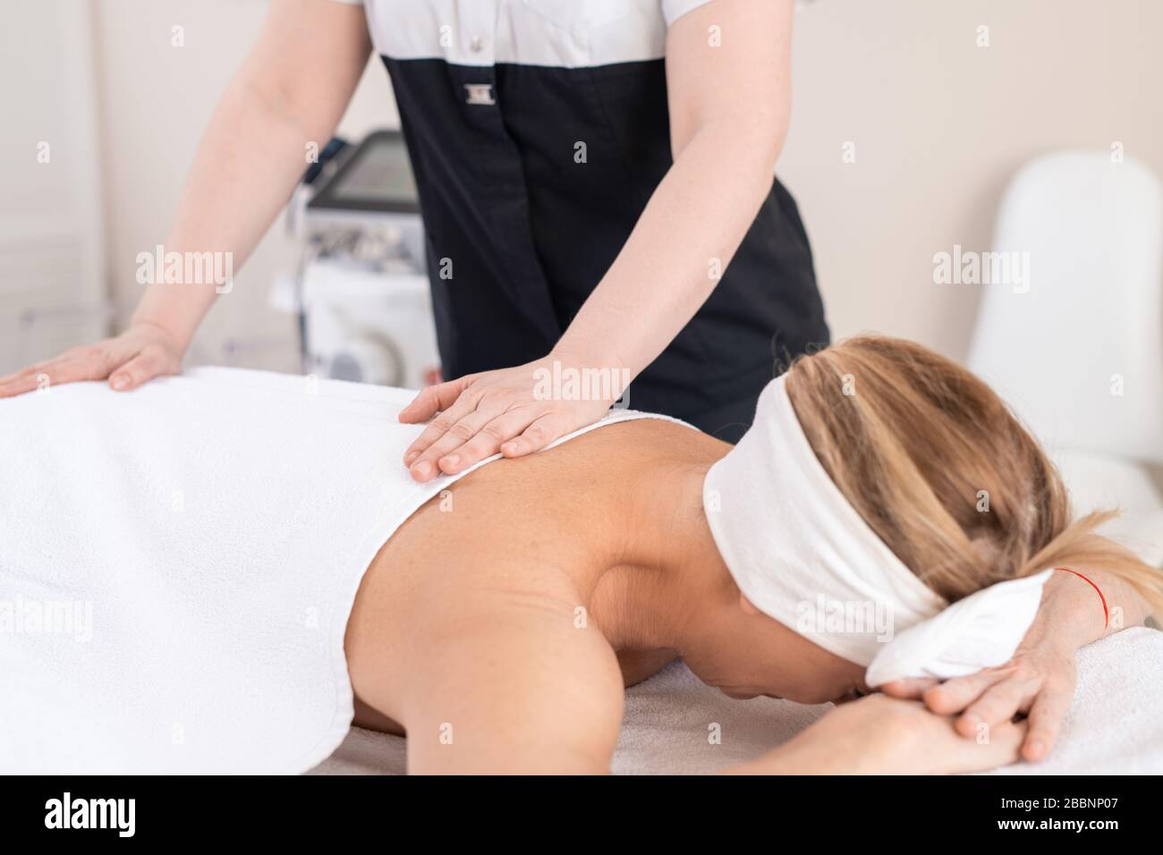 Unerkennbarer Massagetherapeut, der sich hinter der Frau, die unter einem Handtuch liegt und sich mit dem Gesicht an den Händen anlehnt, verwöhnt Stockfoto
