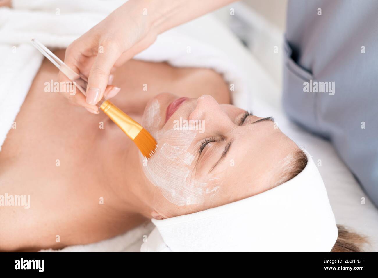 Nicht erkennbarer Kosmetologe, der bei Schönheitsbehandlungen Gesichtsmaske mit Pinsel auf reife Frau anwendet Stockfoto