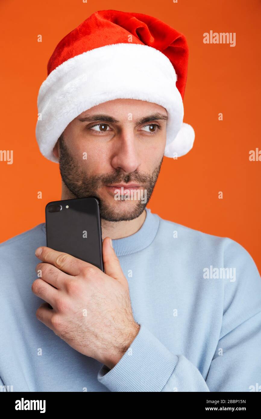 Bild eines ernsthaften jungen Mannes in weihnachtsmann Hut isoliert über orangefarbenem Wandhintergrund mit Handy. Stockfoto