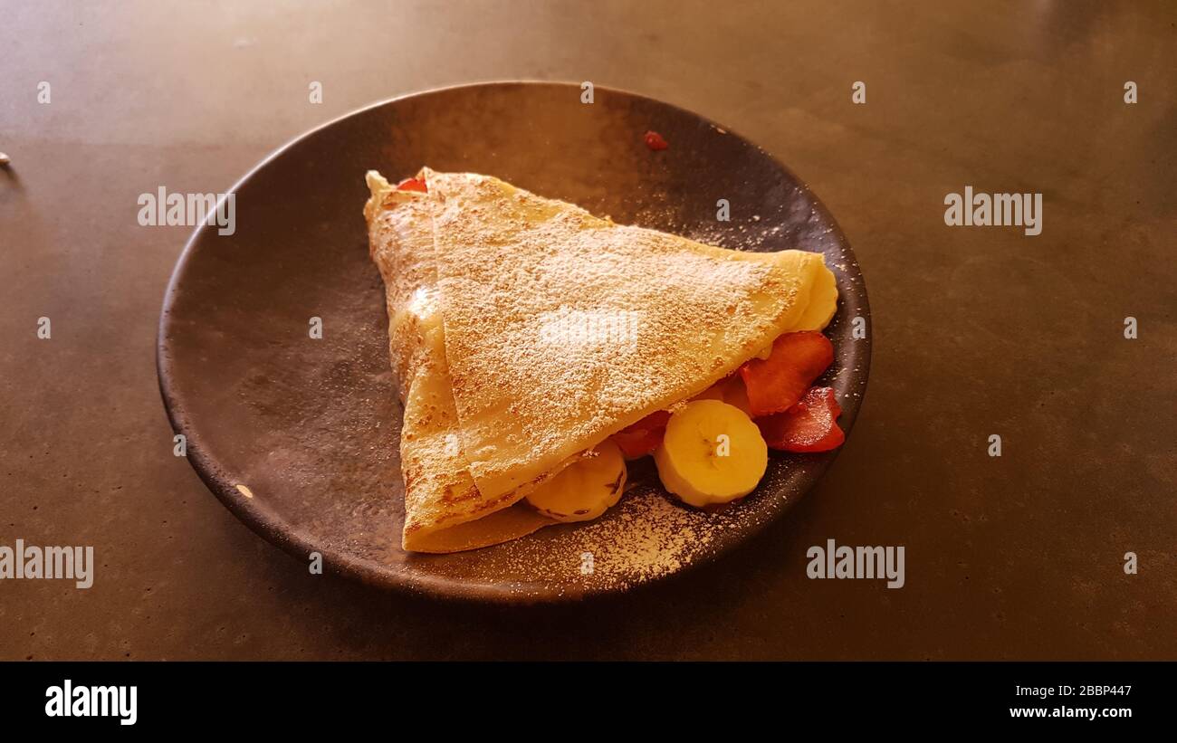 Pfannkuchen/Crepe Nahaufnahme mit Früchten und Konditorzucker wie Banane, Erdbeere auf einem Teller auf dunklem Tisch - hausgemachtes süßes Dessert oder Frühstück Stockfoto