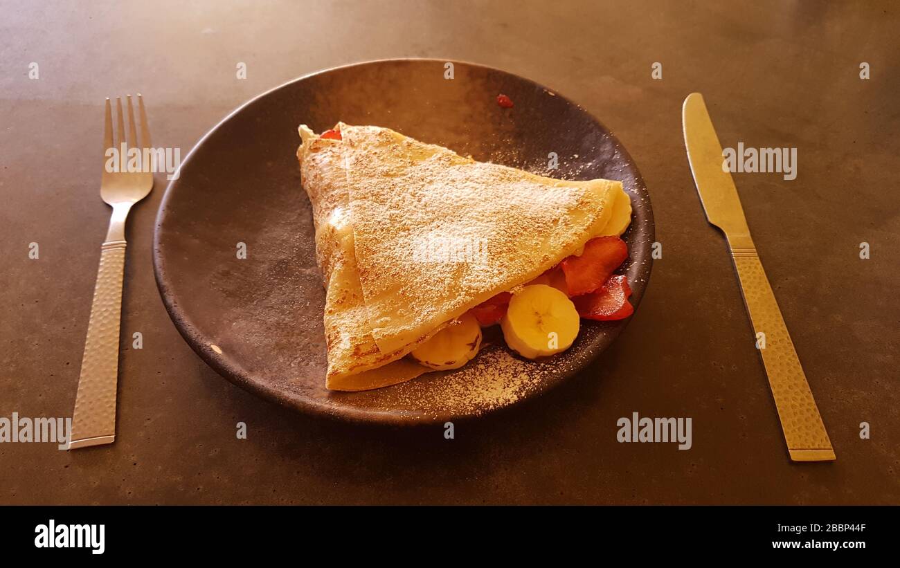 Pfannkuchen/Crepe Nahaufnahme mit Süßigkeiten-Zucker mit Früchten wie Banane, Erdbeere auf einem Teller mit Messer und Gabel auf einem dunklen Tisch - süßes Dessert Stockfoto