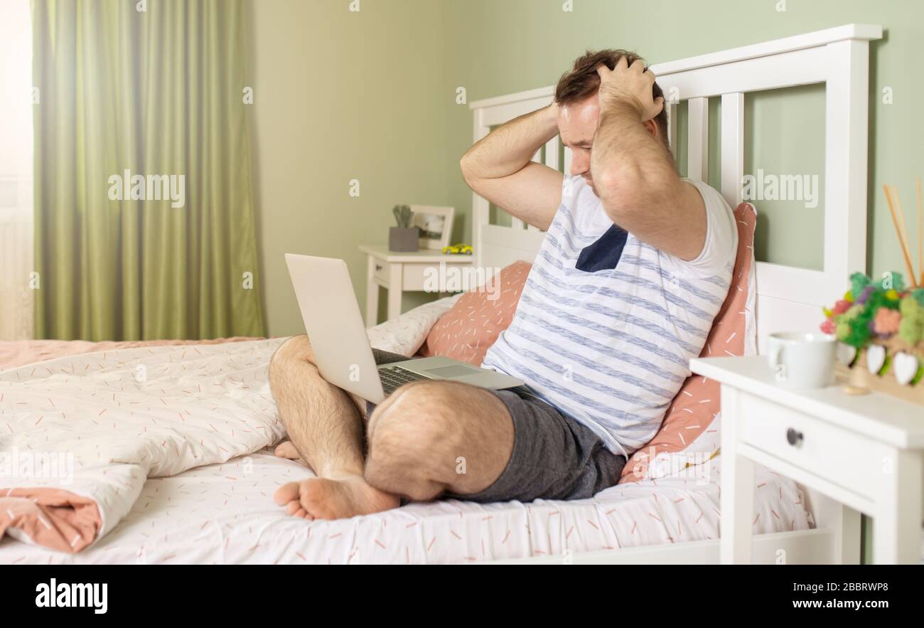 Der Mann mit einem Laptop im Bett erhielt schlechte Nachrichten, er ist verwirrt, hält die Hand am Kopf und beobachtet den Bildschirm in schockierten Augen. Stockfoto