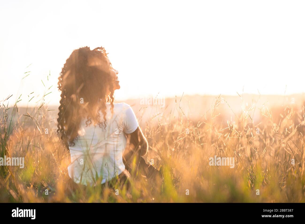 afro-amerikanisches Mädchen auf dem Rücken sitzt bei Sonnenuntergang auf dem Feld und blickt auf den Horizont Stockfoto