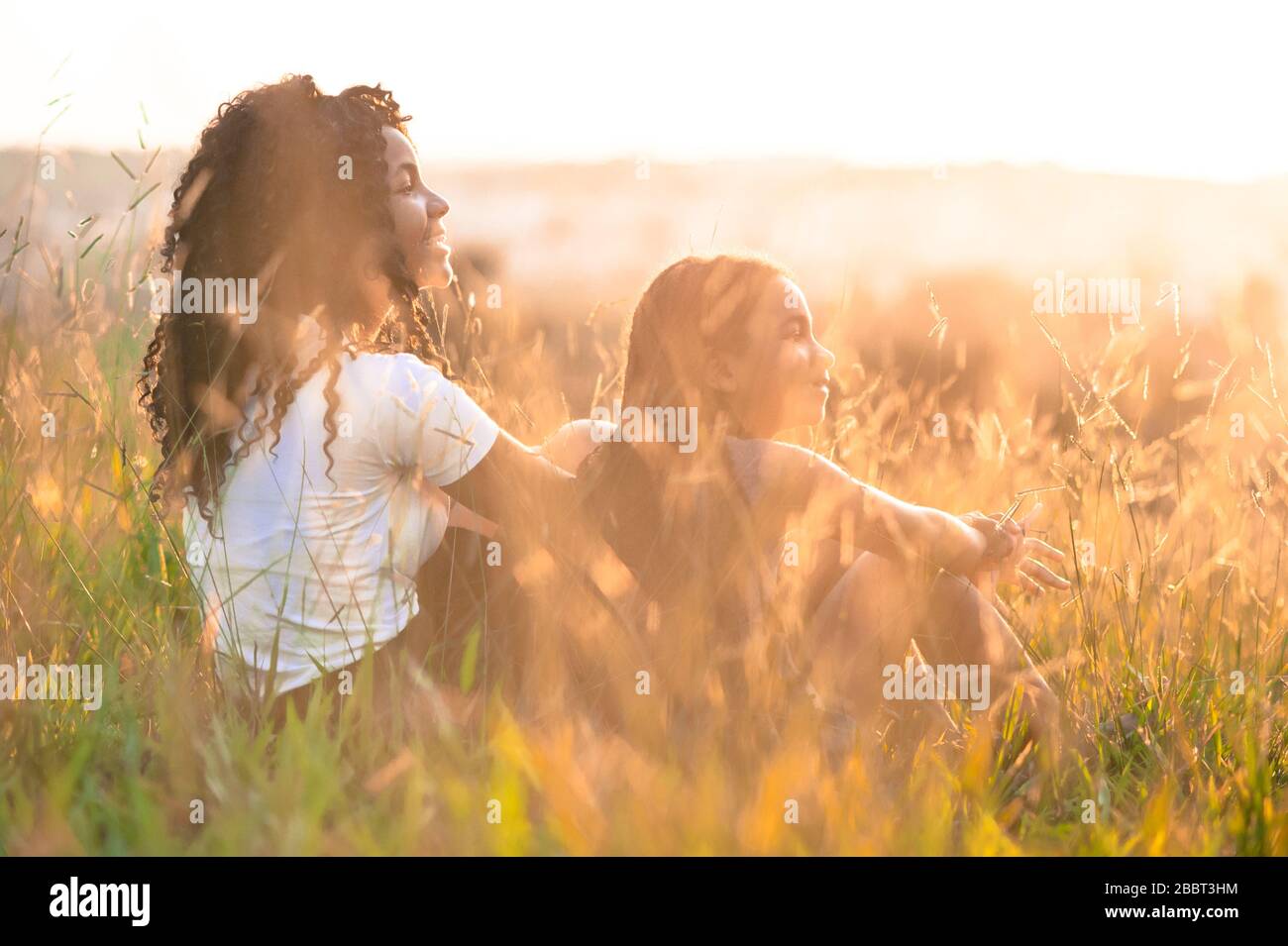 Zwei afro-amerikanische Mädchen sitzen auf dem Feld und blicken auf den Sonnenuntergang mit goldenem Sonnenlicht, das die Szene mit ihrer rechten Seite zur Kurvenscheibe iluminiert Stockfoto