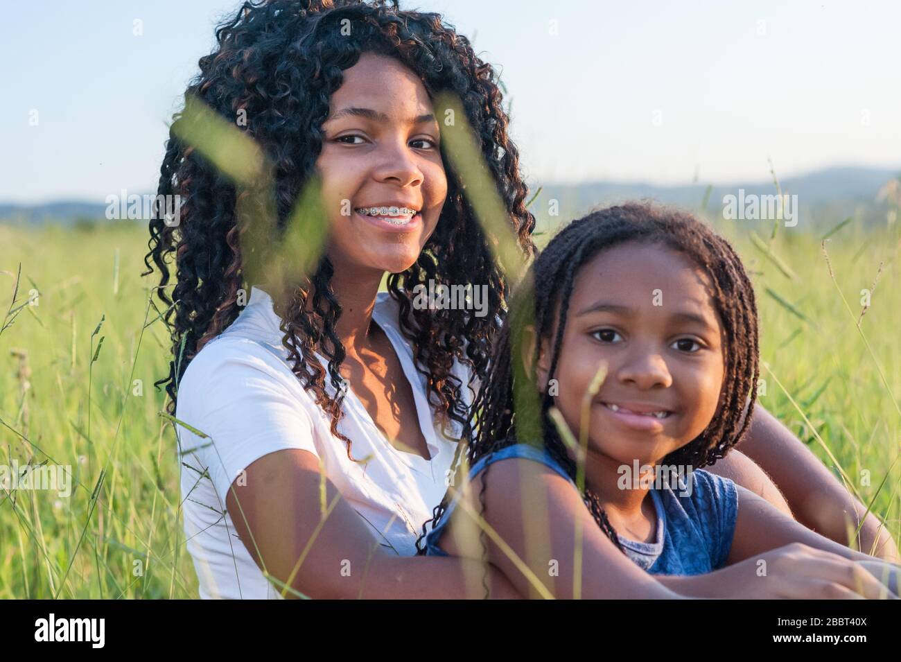Zwei afro-amerikanische Mädchen lächelten auf dem Gras und schauten auf die Kamera. Beide haben lange schwarze Haare. Stockfoto