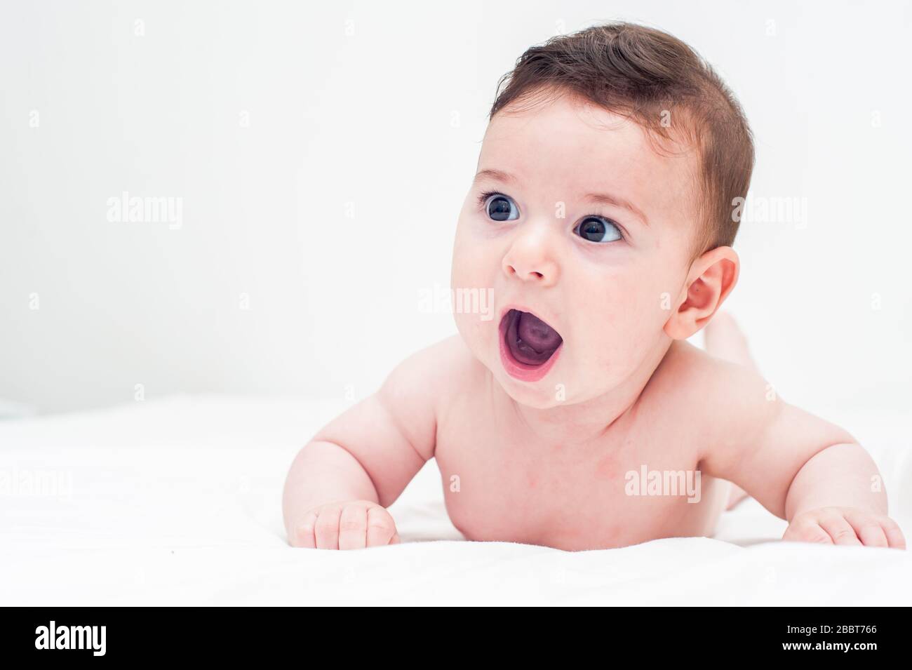 Süßes Baby liegt auf dem Bauch im Bett auf weißem Hintergrund. Sein Mund ist geöffnet. Bild mit großem Kopierraum. Stockfoto