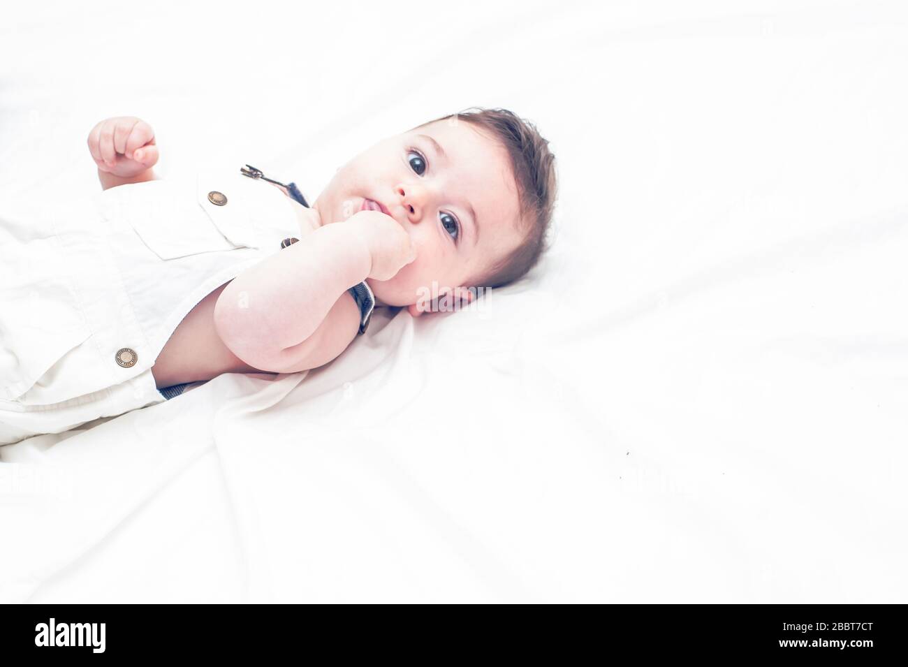 Süßes Baby in weißen Overalls, das auf einem weißen Blatt liegt. Seine linke Hand liegt auf dem Mund. Stockfoto