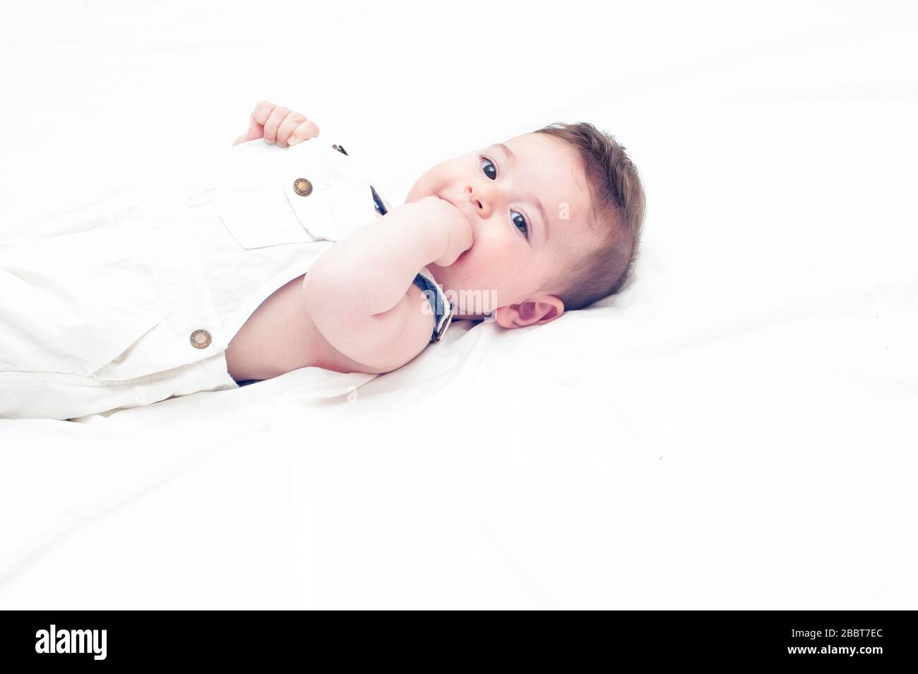 Süßes Baby in weißen Overalls, das auf einem weißen Blatt liegt. Seine linke Hand liegt auf dem Mund. Stockfoto