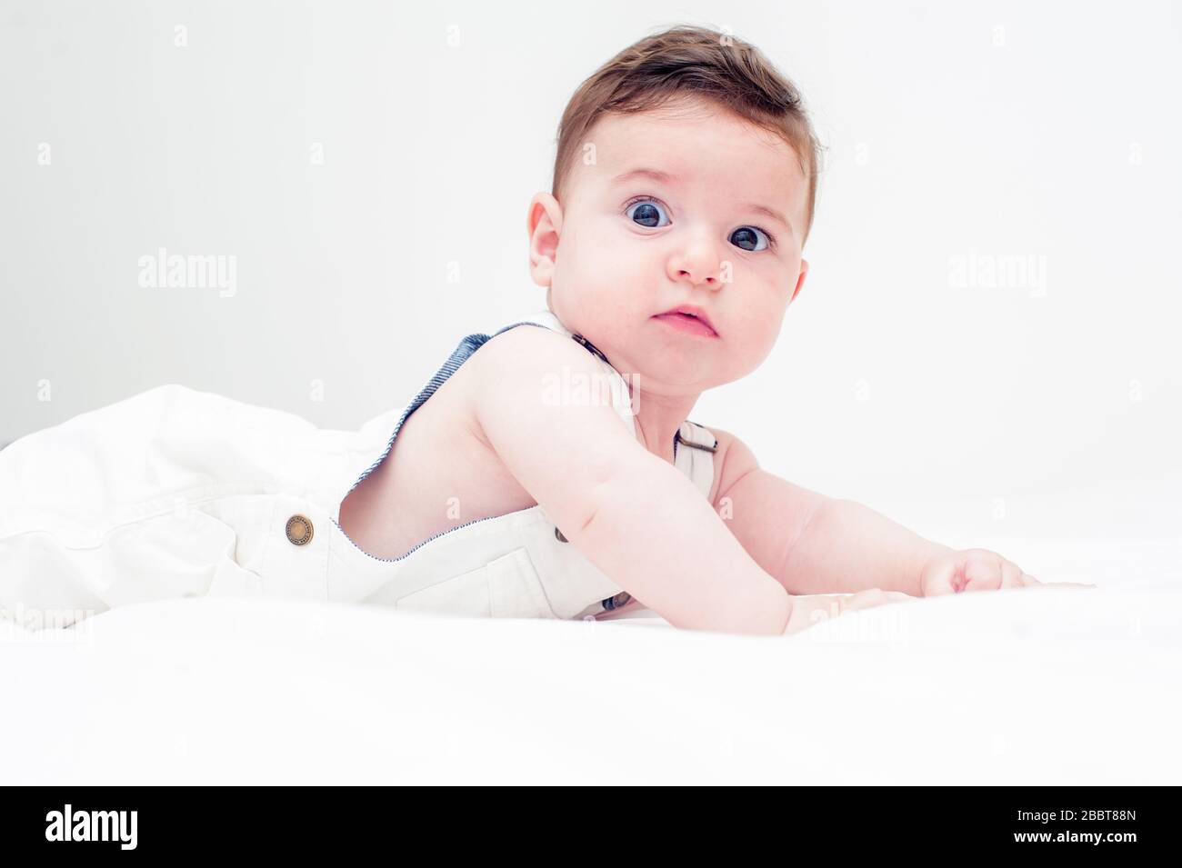 Schönes Baby, das in weißen Overalls auf dem Bauch in einem weißen Blatt und einem weißen Hintergrund auf die Kamenra blickt. Stockfoto