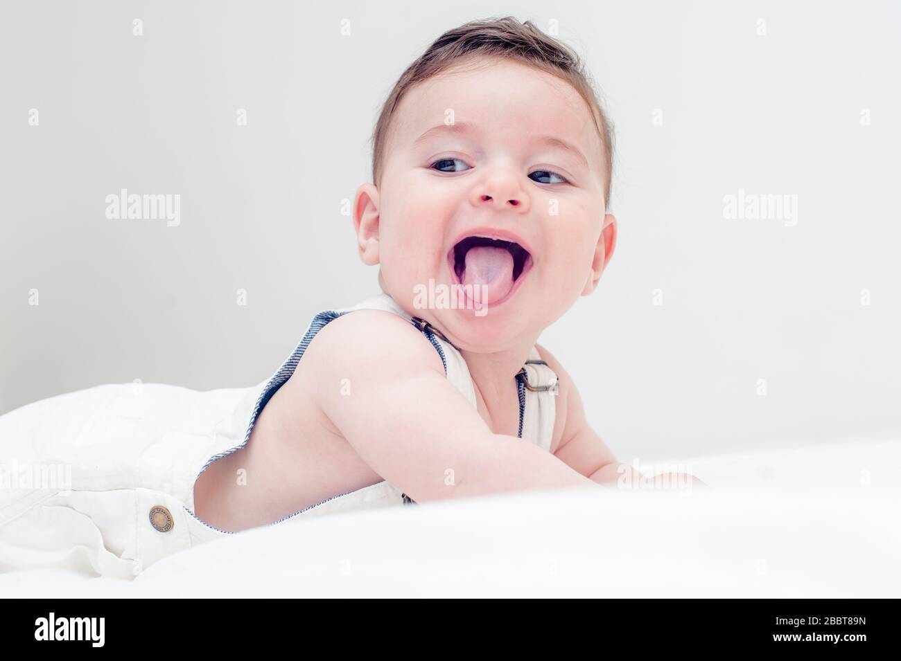 Süßes Baby lächelt mit seiner Zunge in weißen Overalls, die in einem weißen Blatt auf dem Bauch liegen. Stockfoto