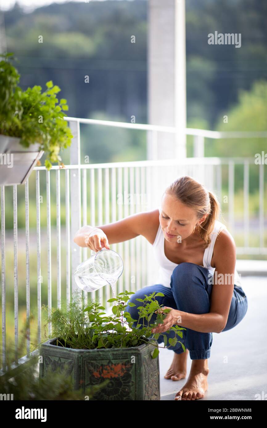 Hübsche, junge Frau, die Kräuter wässert, die sie auf ihrem Balkon wächst. Stockfoto