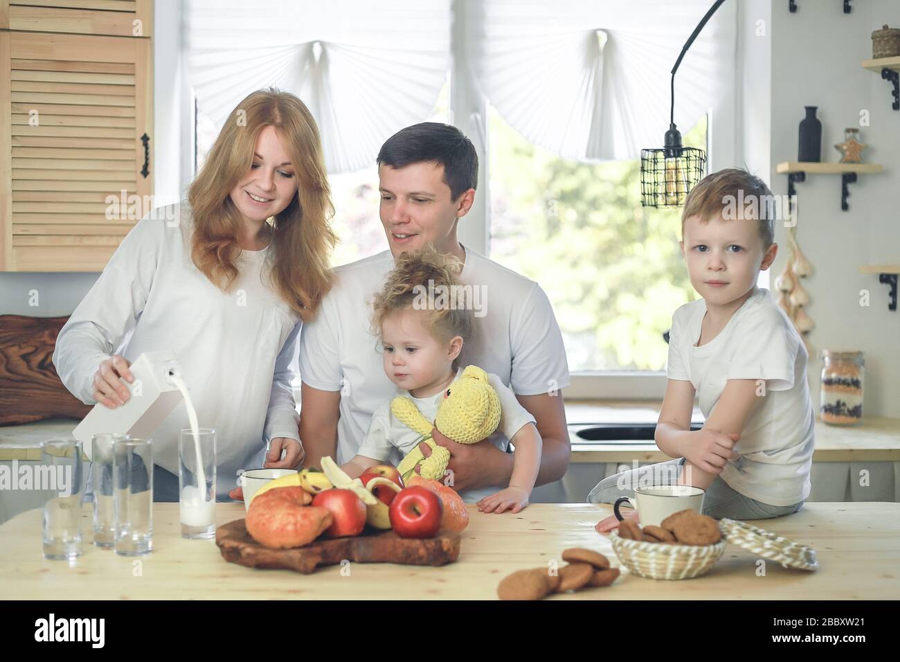 Glückliche Familienmutter, Vater, Tochter und Sohn, die zu Hause frühstücken. Mutter gießt Milch in ein Glas. Vitaminfrühstück im hellen Küchenraum Stockfoto