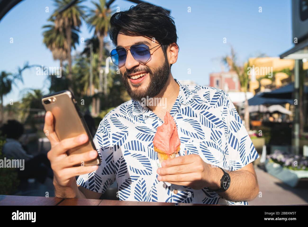 Porträt des jungen Mannes, der bei schönem Wetter eine Eiscreme isst und sein Handy im Freien benutzt. Stockfoto