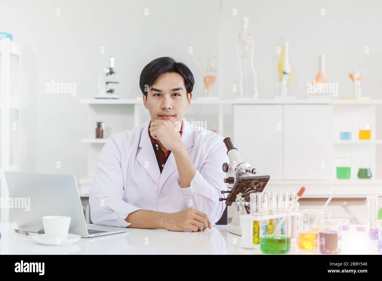 Junger, gutaussehender Wissenschaftler, der im Laborarbeitstisch sitzt, Asian Race, der mit der Kamera Hand auf das Kinn blickt Stockfoto