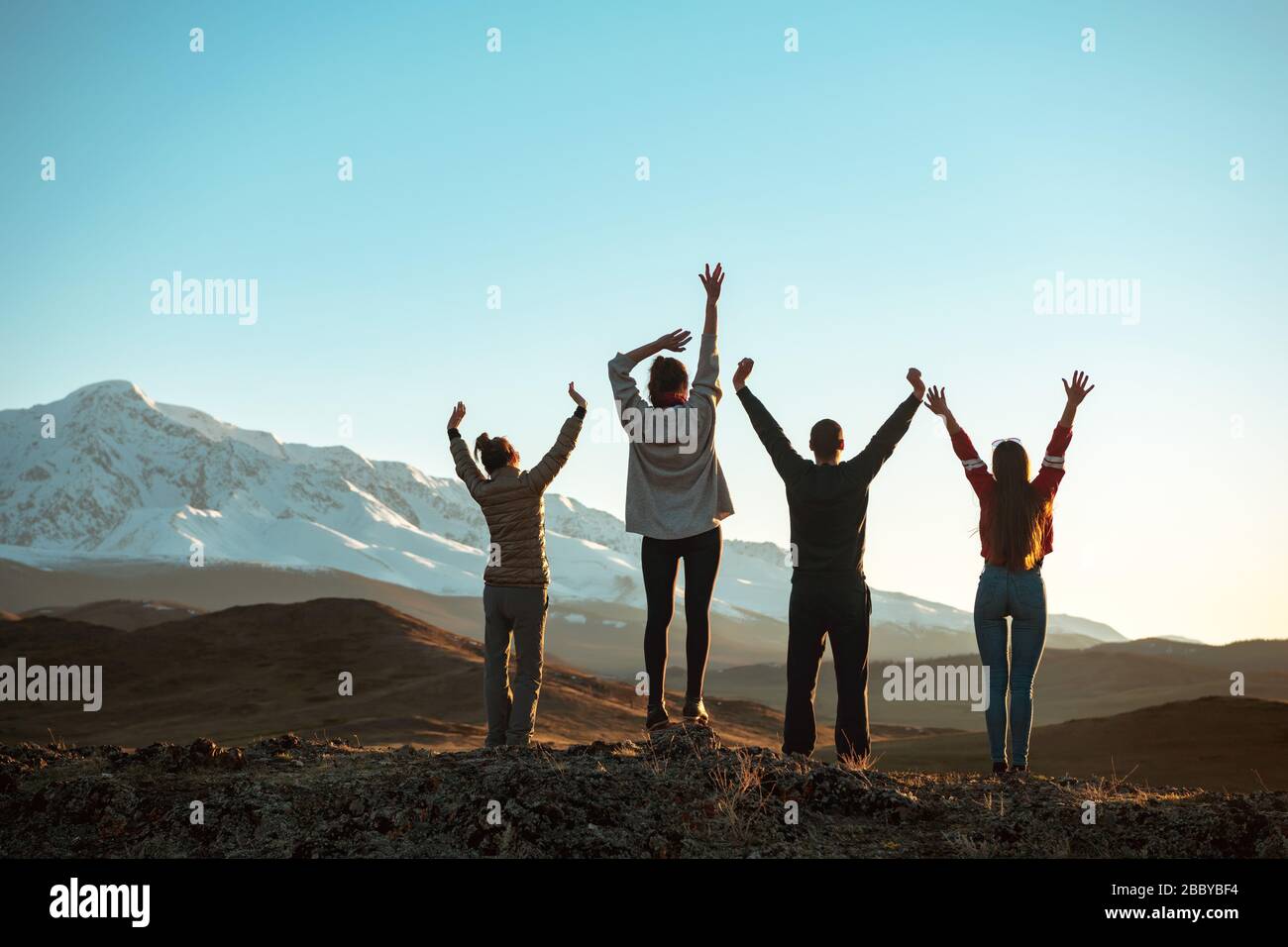 Vier glückliche Freunde stehen mit erhobenen Armen und blicken auf Berge und Himmel bei Sonnenuntergang. Nicht anerkannte Völker Stockfoto