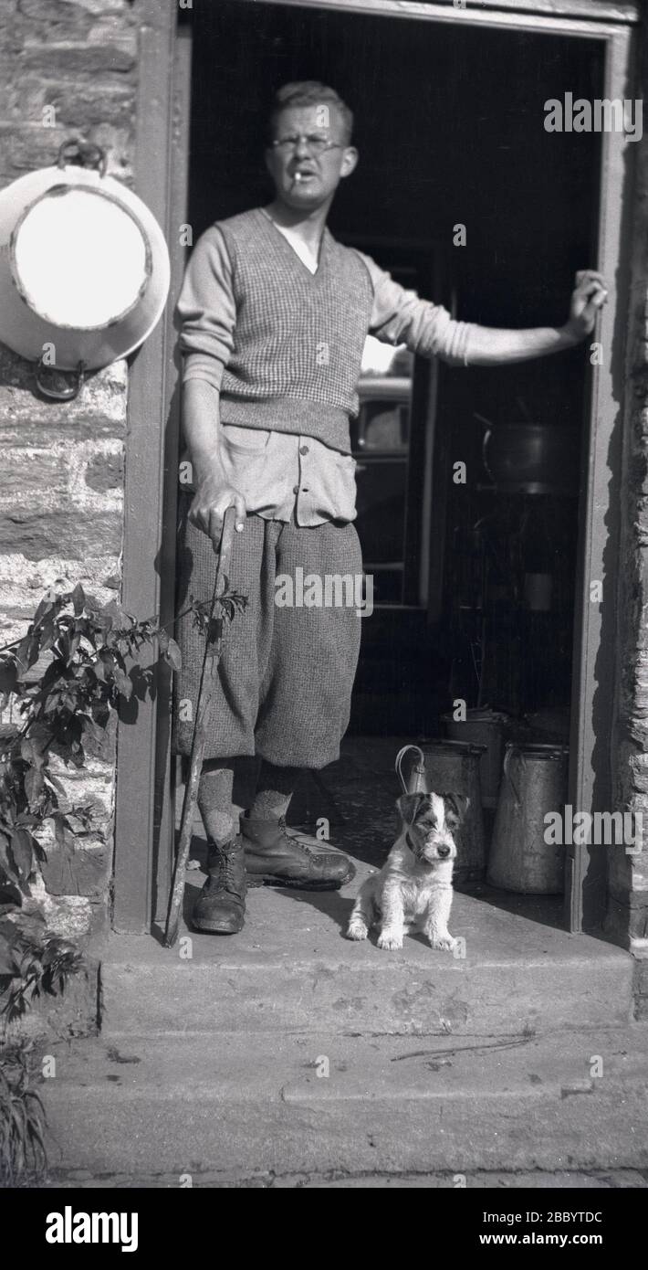 Dreißiger Jahre, ein Landarbeiter in der Kleidung, die für die Epoche typisch ist, einschließlich Mehl und Zigarette im Mund, mit Haustierhund, der in der Tür seines Cottages, Devon, England, steht. Eine Reihe von Milchfässern ist im Eingang zu sehen, während außen an der Wand neben der Tür eine Melkschale aus Metall hängt. Stockfoto