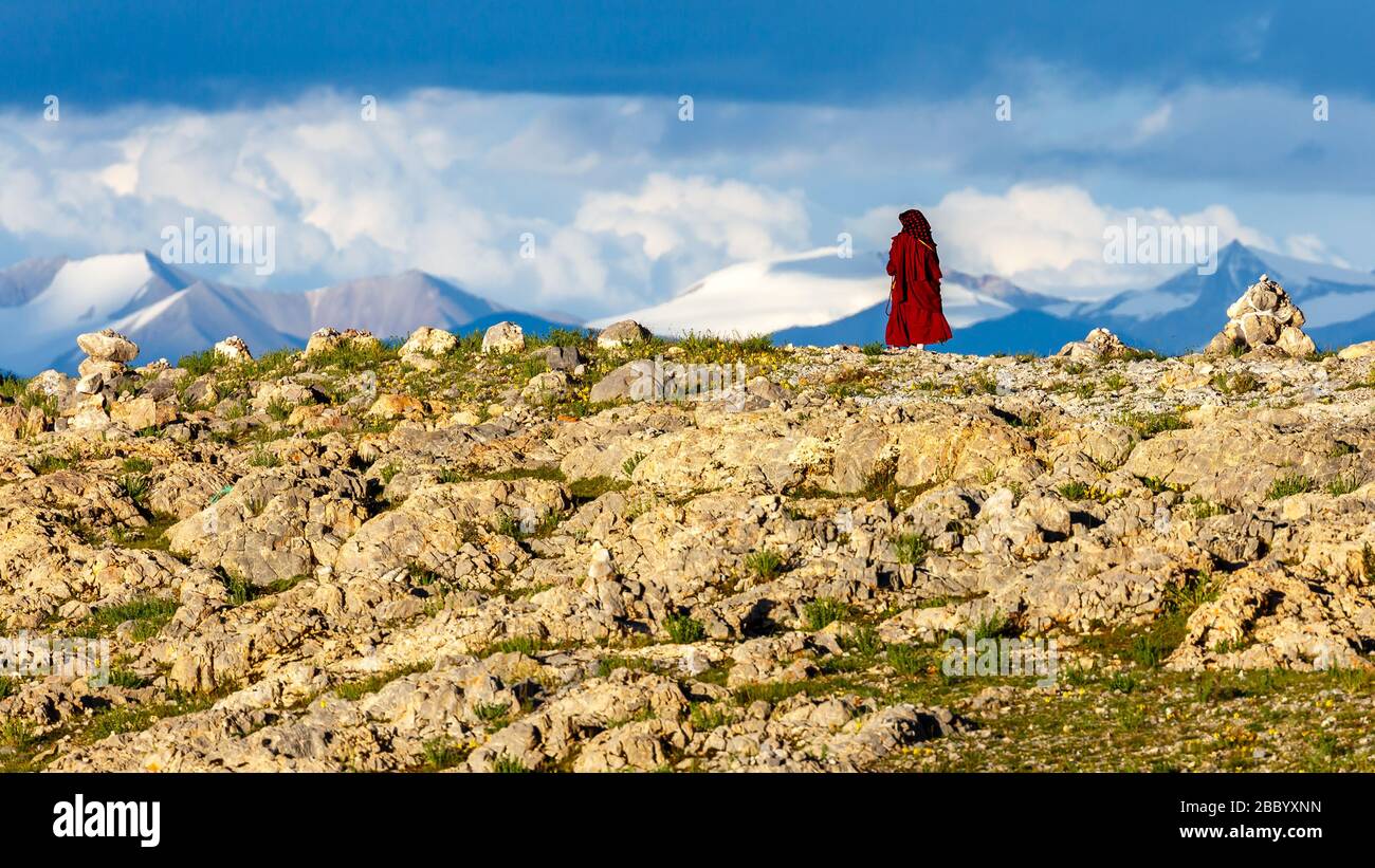 Buddhistischer Pilger in roter Robe, der am Nam Tso umherwandert. Für tibeter ist das Gebiet um den See heilig. Mit Wolken und schneebedeckten Bergen. Stockfoto