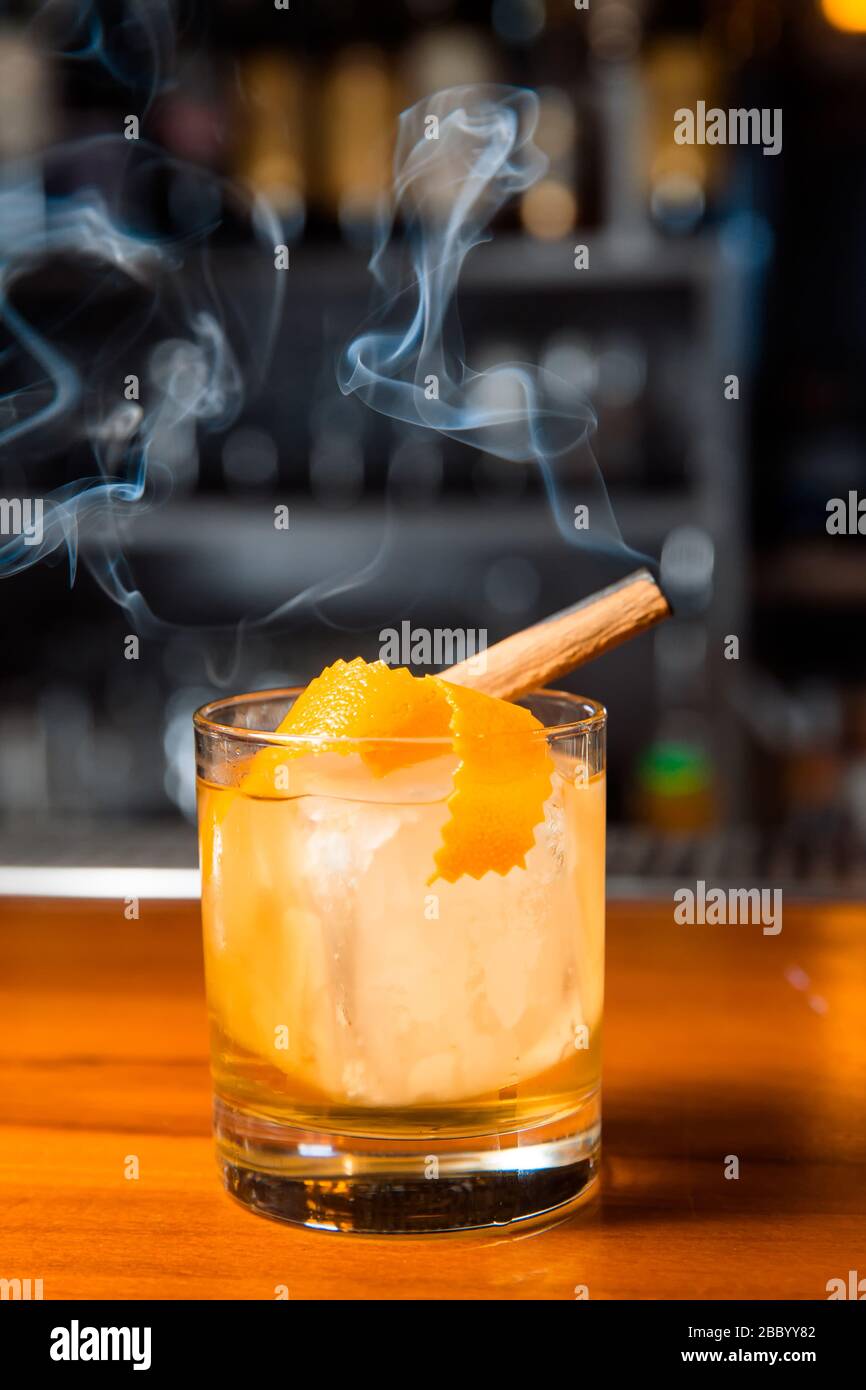 Orangencocktail mit Eiswürfel und Orangenschale. Im Glas ist ein Zimtstab, aus dem Rauch entsteht. Stockfoto