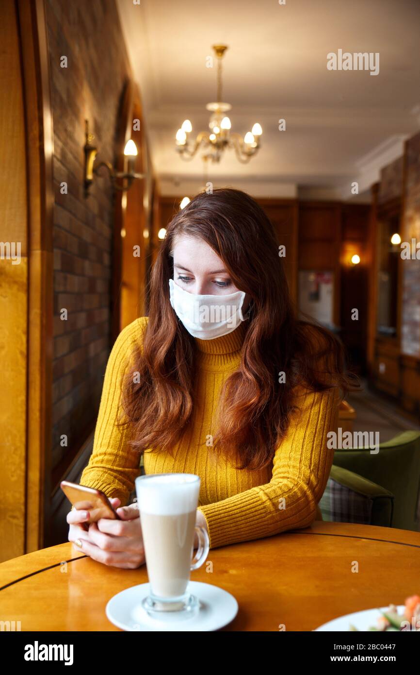 Junge kaukasische Frau, die Gesichtsschutzmaske trägt und Nachrichten auf ihrem Handy in der Halle liest. Stockfoto