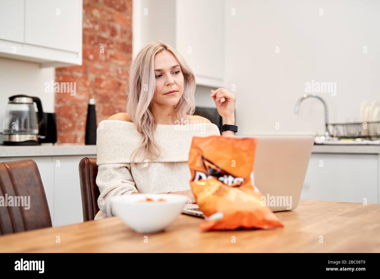 Eine einzelne blonde kaukasische Frau, die an einem Tisch sitzt und einen Laptop mit einer Tüte mit tangarem Käse Doritos daneben betrachtet Stockfoto