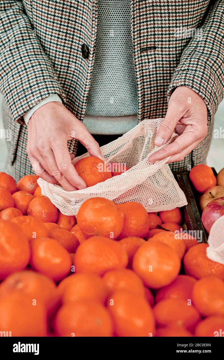 Nahaufnahme eines Mannes, der in einem Greengrocker einkauft und einige Mandarinorangen in einen wiederverwendbaren Textil-Netzbeutel einbringt, um die Kunststoffverschmutzung zu verringern Stockfoto