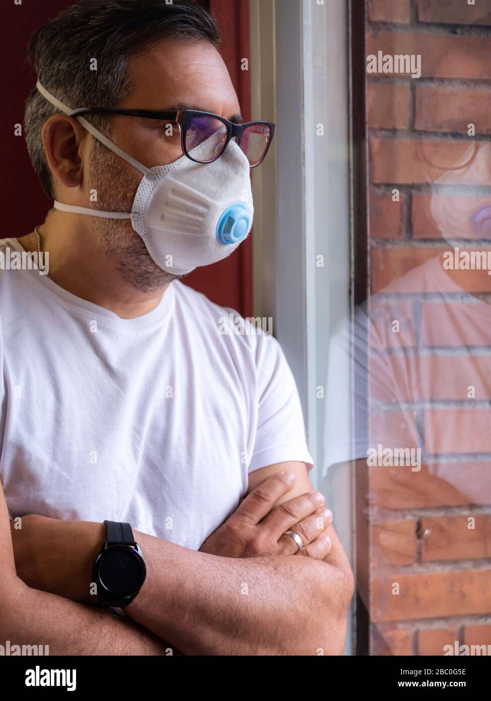 Ein Mann mit einer schützenden medizinischen Maske wartet auf das Ende der Kovid-19-Notquarantäne, hinter dem Glas seines Heimatfensters. Stockfoto