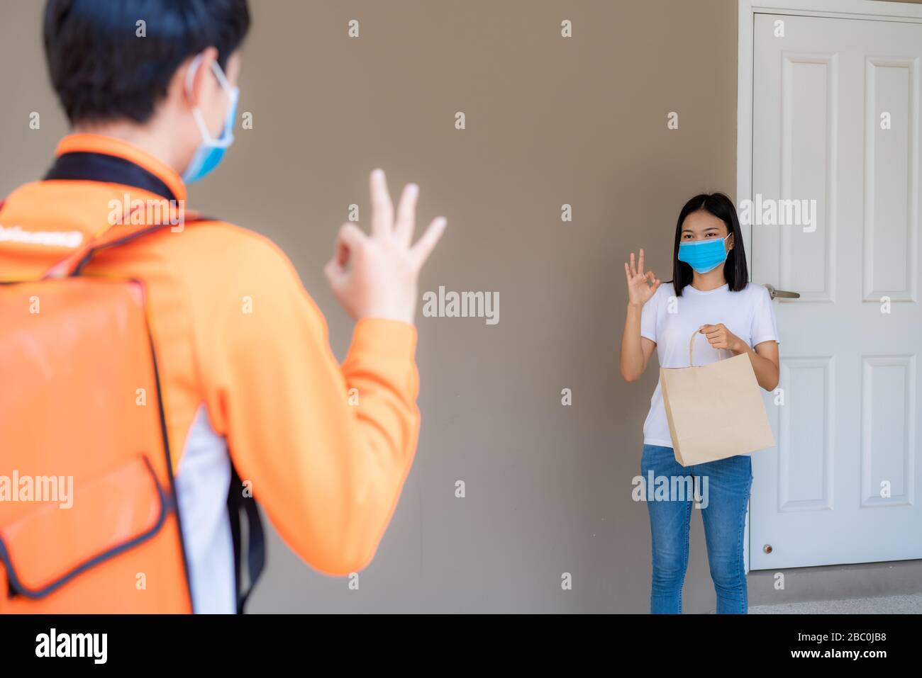 Asian Woman holt Lieferlebensmittelbeutel vom Türknopf und OK-Schild für kontaktlose oder Kontakt frei von Lieferfahrern mit Fahrrad im Vorderhaus für s Stockfoto