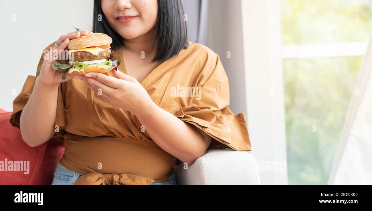 Hungrig übergewichtige Frau und lächelnd Hamburger und sitzen im Wohnzimmer, sie sehr glücklich und genießen Sie Fast Food zu essen. Konzept der binge eati Stockfoto