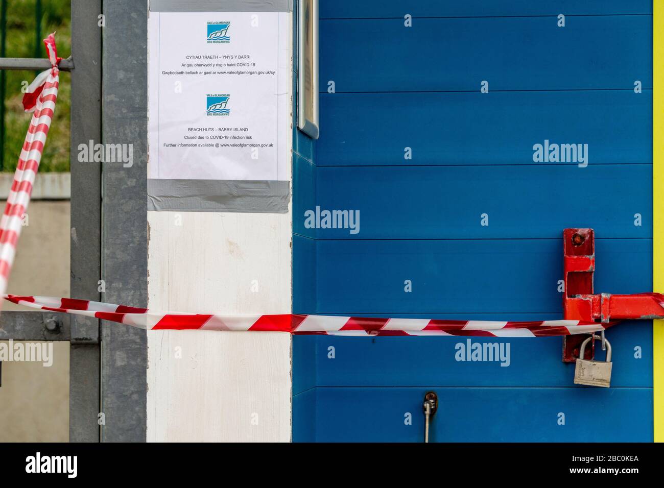 Nahaufnahme der mit einem Vorhängeschloss abgeschlossenen Strandhütentür mit Covid-19-Hinweis und rot-weiß gestreiftem Band, das den Zugang einschränkt. Stockfoto