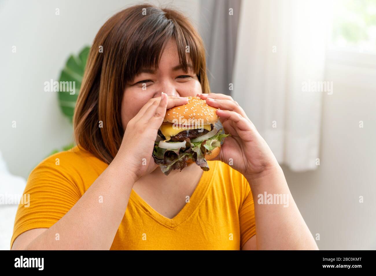 Hungrige, übergewichtige junge asiatin, die Hamburger hält, ihre hungrigen und überernährten, gluttonigen und bingen Essen. Ihr Lebensstil isst fast Food a Stockfoto