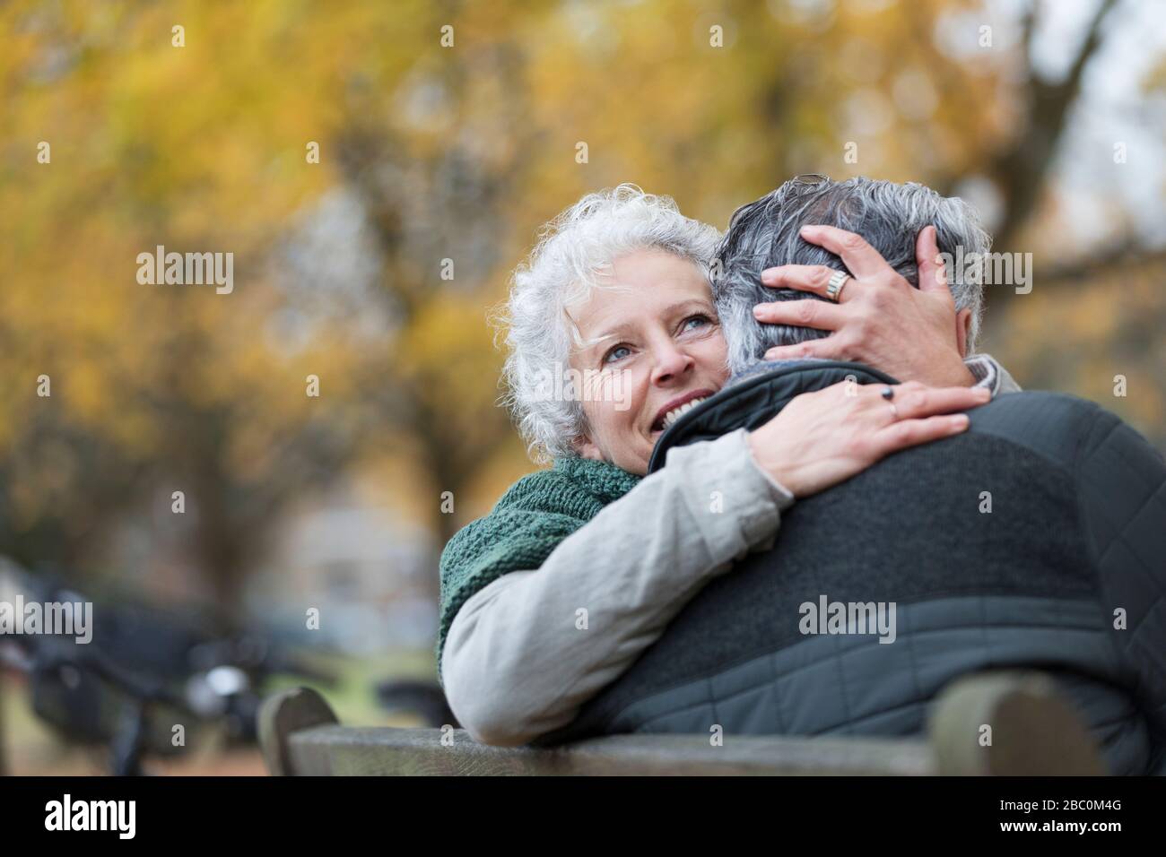 Lächelndes, liebevolles Senioren-Paar, das sich im Herbstpark auf der Bank umarmt Stockfoto