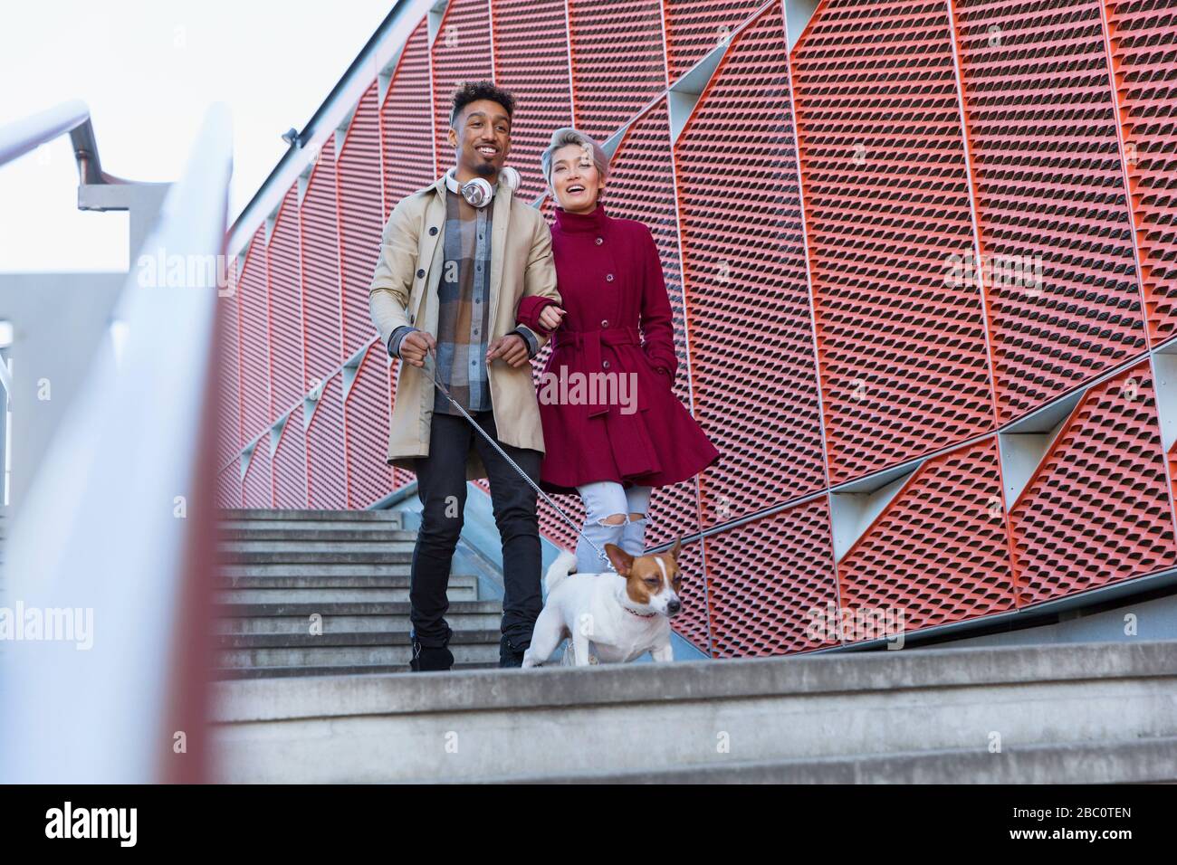 Lächelndes junges Paar, das mit dem Hund die urbanen, modernen Treppen hinuntergeht Stockfoto